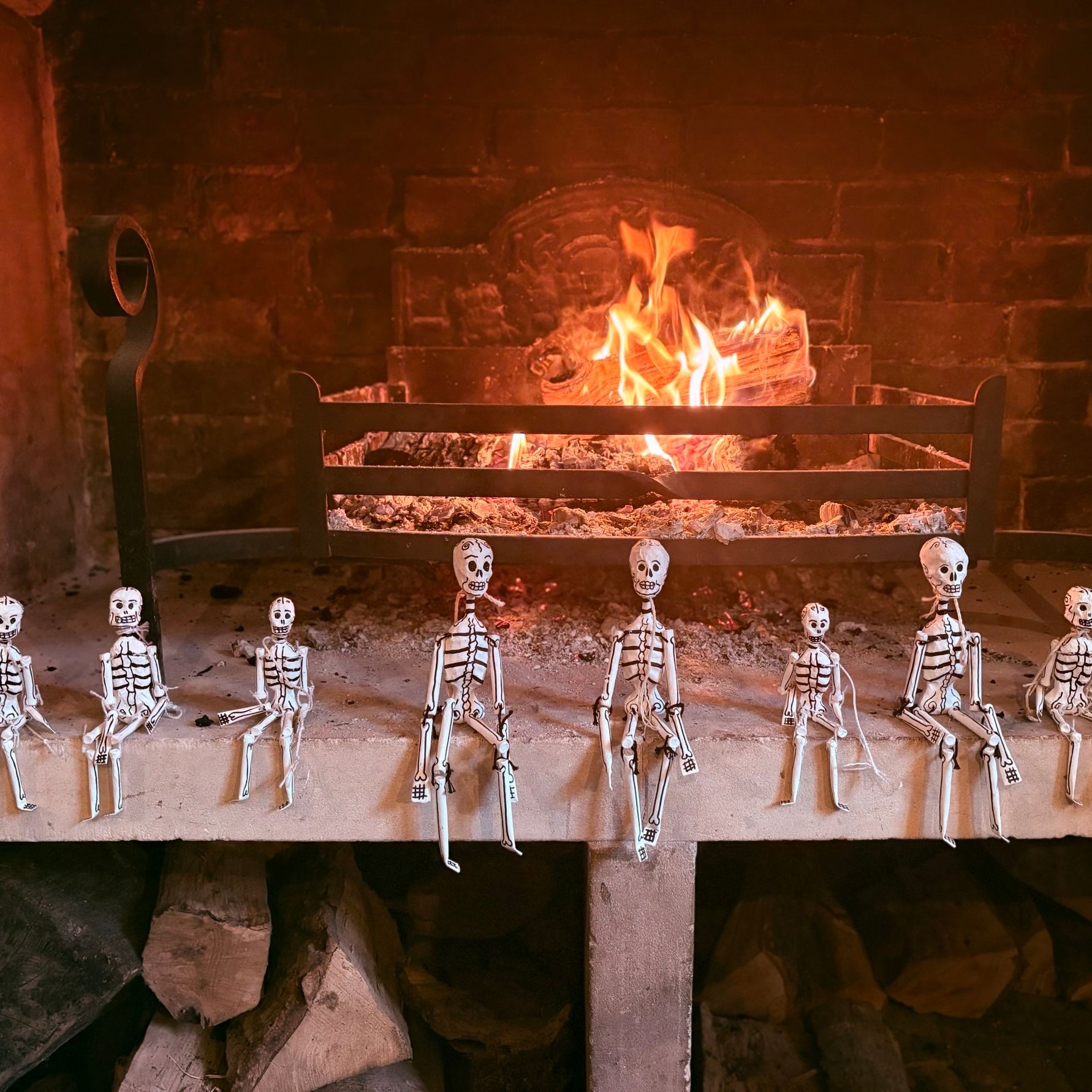 Paper skeleton decorations lined up in front of a burning log fire in a brick fireplace