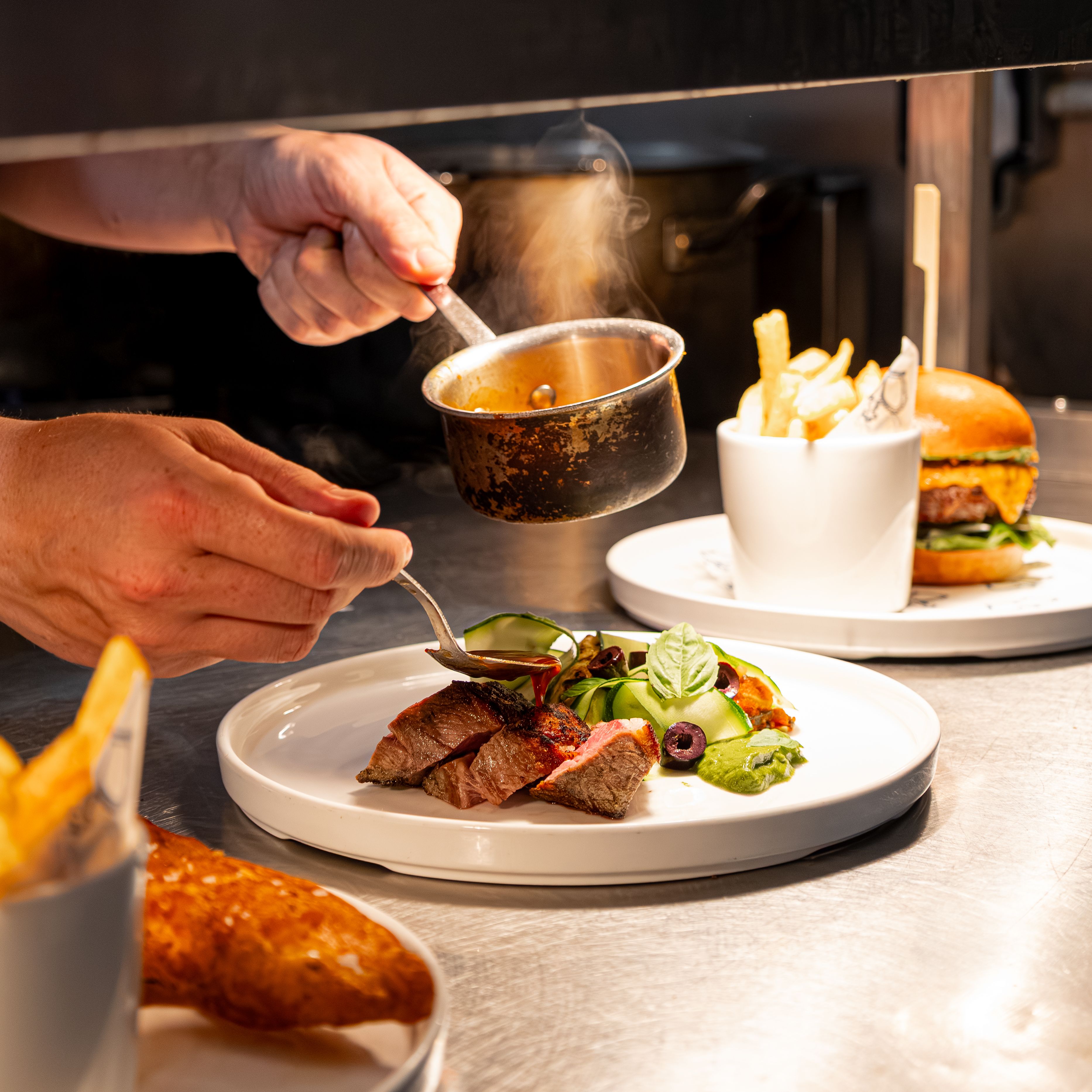 Chef plating steak dish with sauce next to burger and fries in a restaurant kitchen