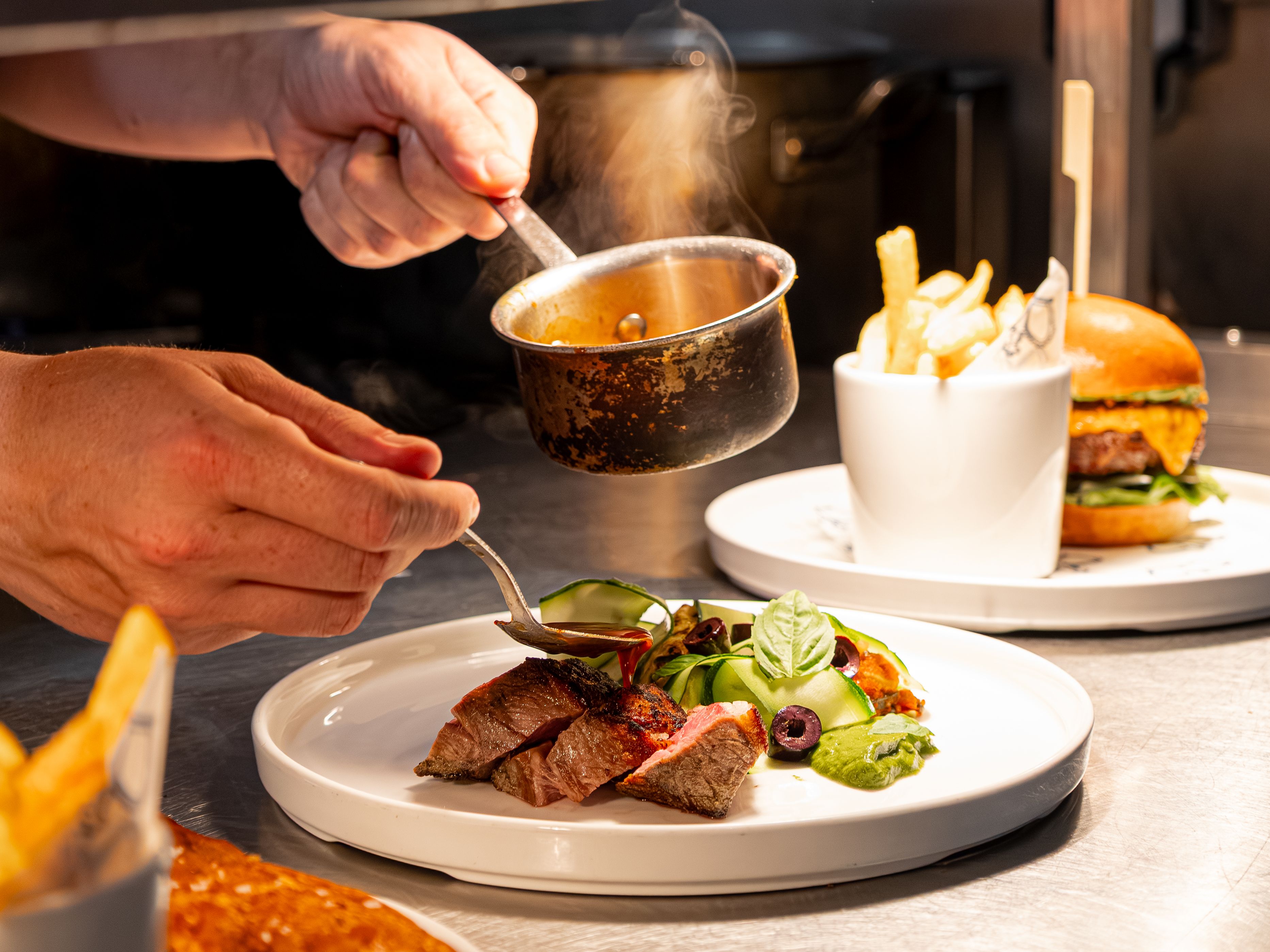 Chef plating steak dish with sauce next to burger and fries in a restaurant kitchen