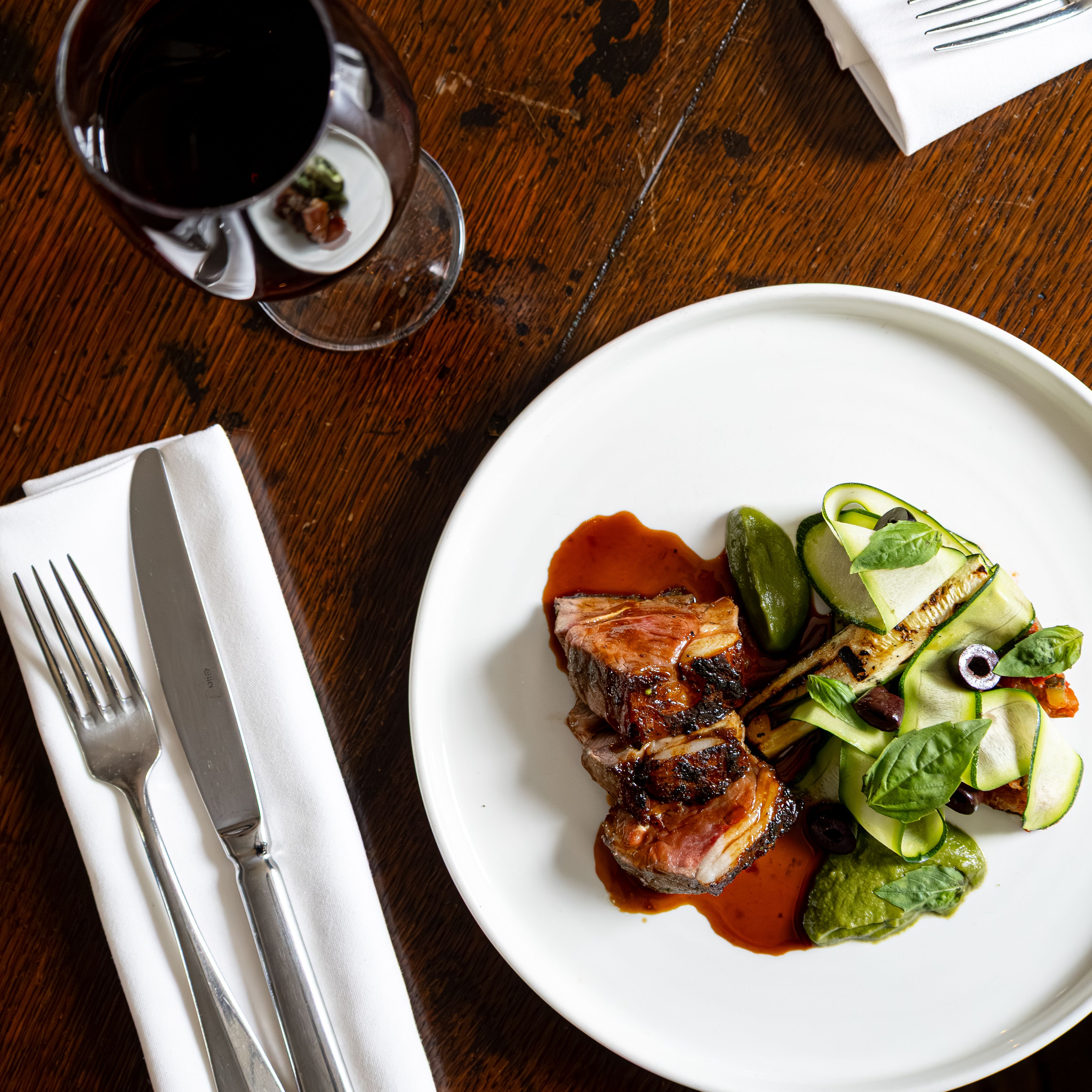Gourmet meal with sliced meat, vegetables, and sauce on a white plate, next to a glass of red wine and cutlery on a rustic wooden table.