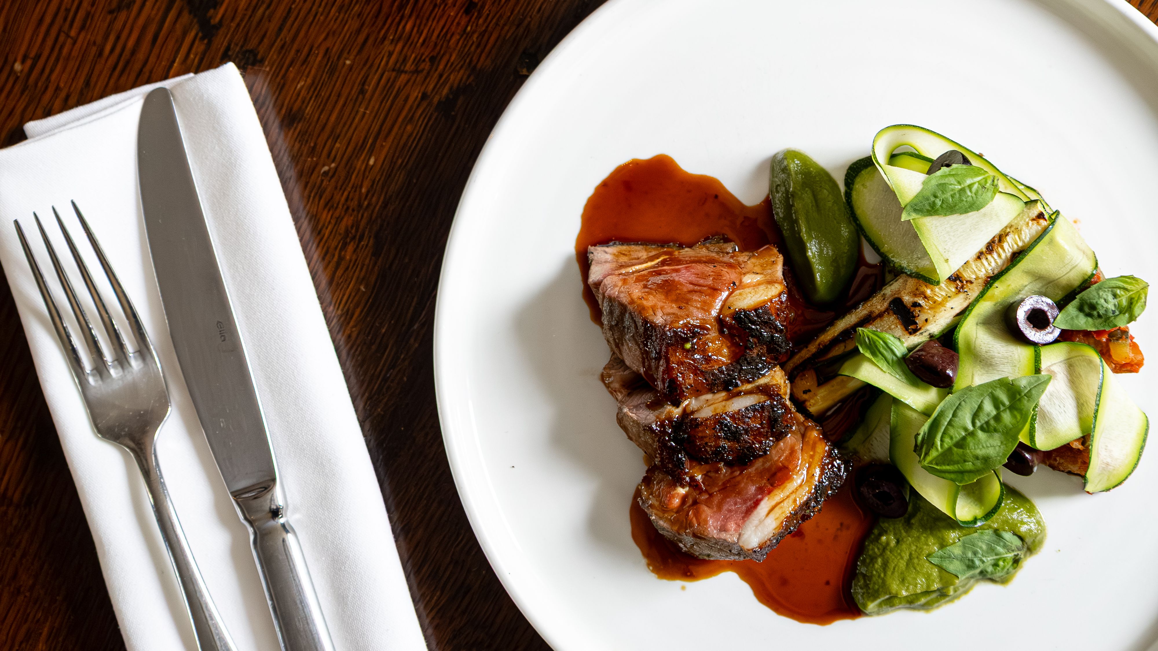 Gourmet meal with sliced meat, vegetables, and sauce on a white plate, next to a glass of red wine and cutlery on a rustic wooden table.