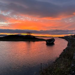 A vibrant sunset near the beaches in Sligo, with an old boat partially submerged in a calm river. Orange and purple hues fill the sky, reflecting softly on the water.