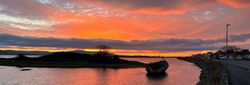 A vibrant sunset near the beaches in Sligo, with an old boat partially submerged in a calm river. Orange and purple hues fill the sky, reflecting softly on the water.