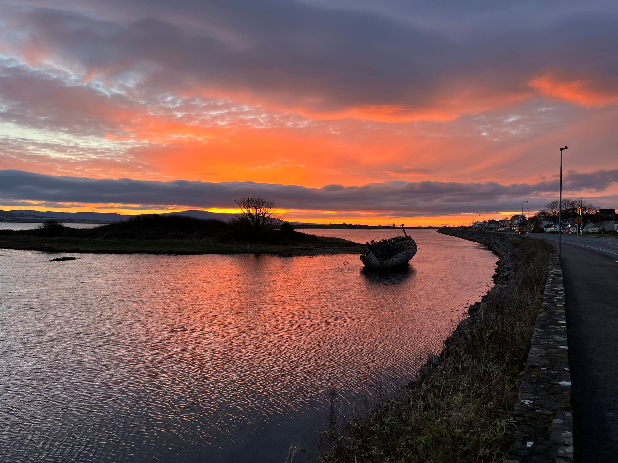 A vibrant sunset near the beaches in Sligo, with an old boat partially submerged in a calm river. Orange and purple hues fill the sky, reflecting softly on the water.