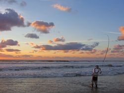 An angler stands at the edge of the water, casting a line into the waves during a peaceful sunset at the beaches in Sligo. The sky glows in soft hues of orange and blue.