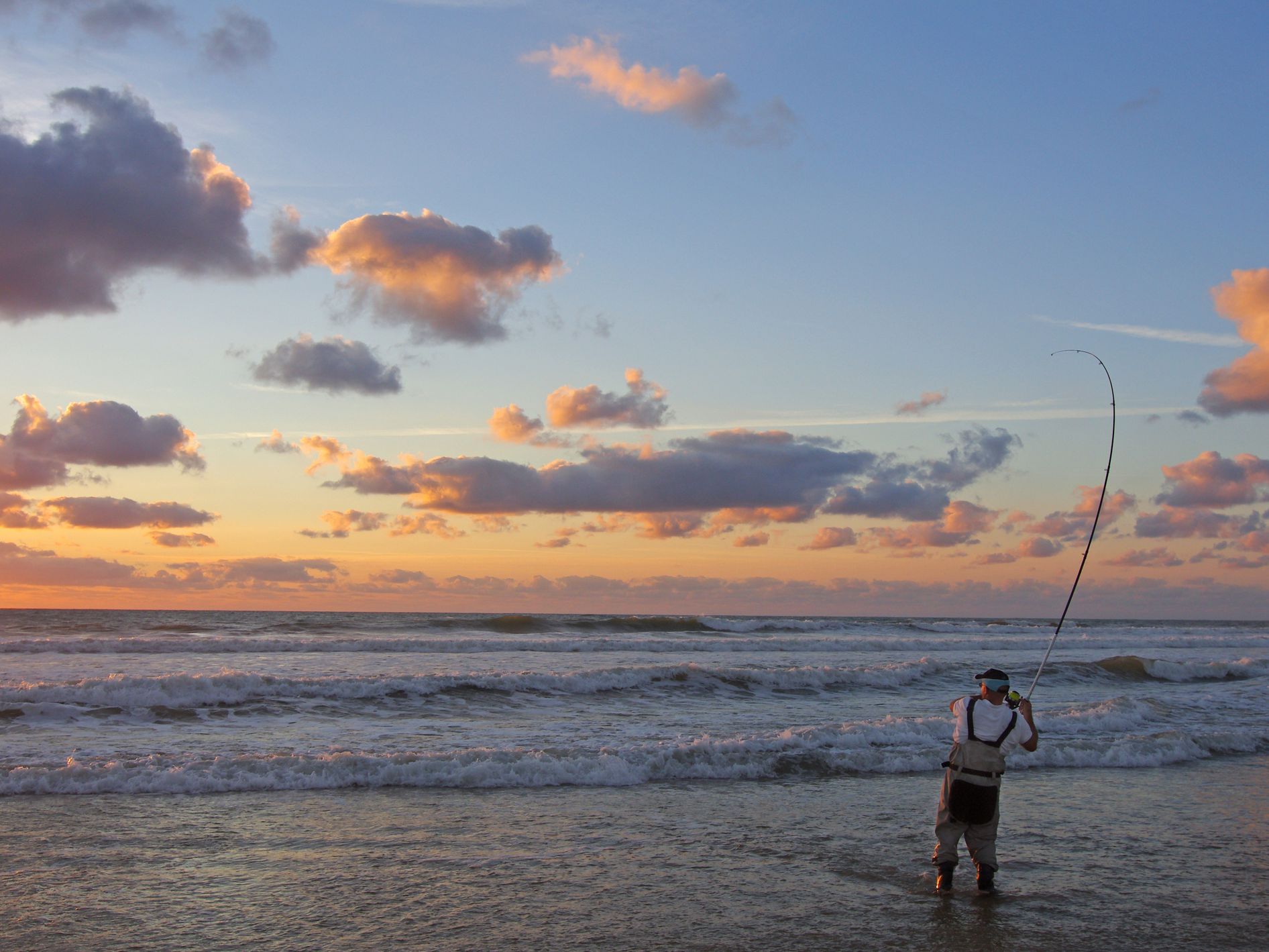 An angler stands at the edge of the water, casting a line into the waves during a peaceful sunset at the beaches in Sligo. The sky glows in soft hues of orange and blue.