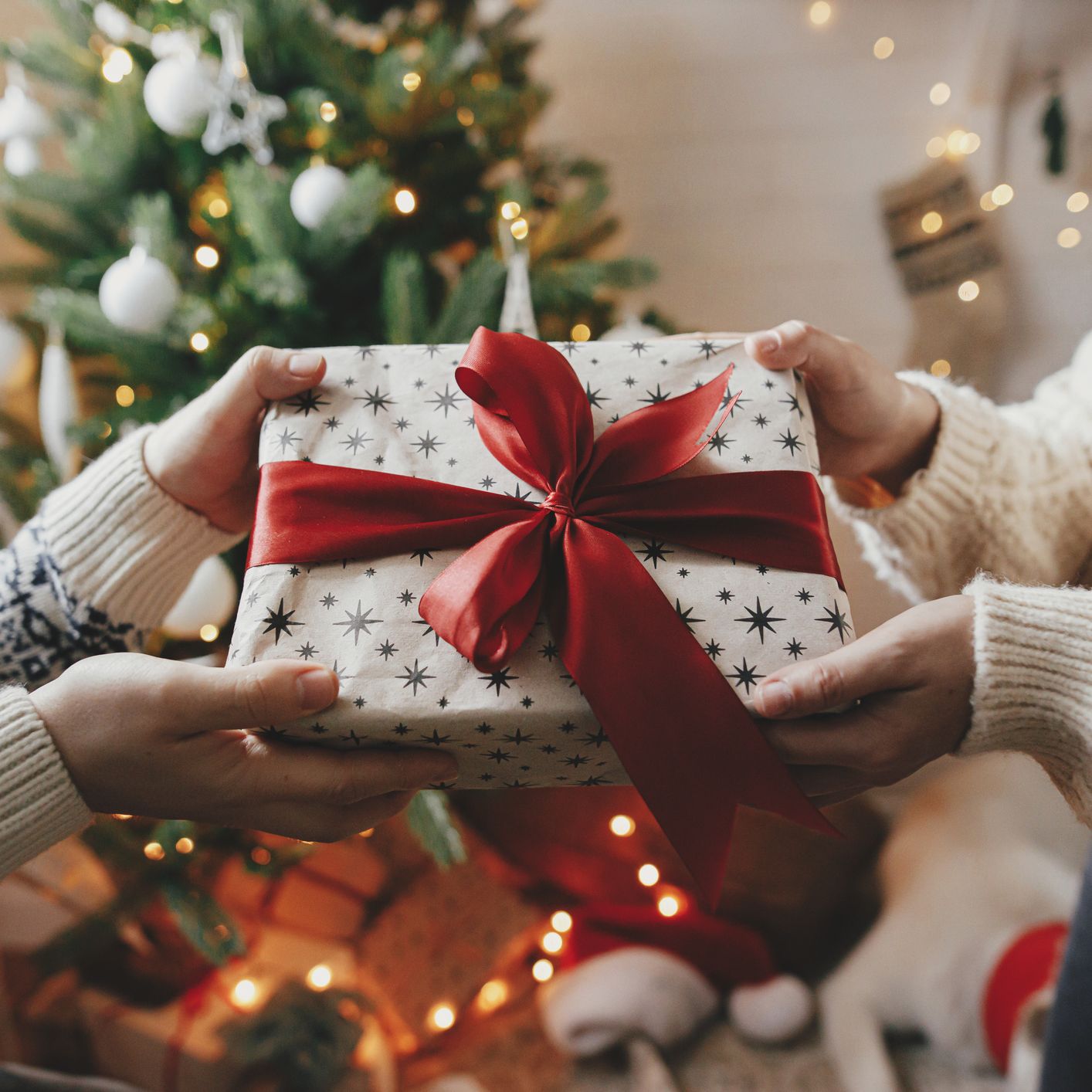 Two people exchange a beautifully wrapped present with a red ribbon in front of a twinkling Christmas tree, inspiring thoughtful Christmas gift ideas.