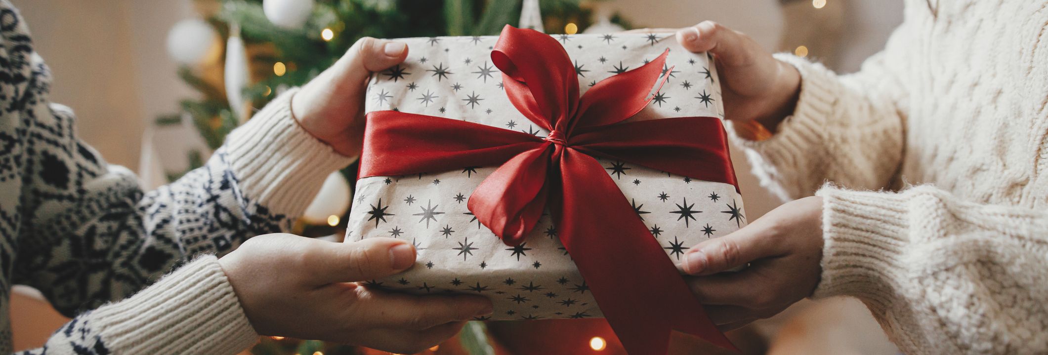Two people exchange a beautifully wrapped present with a red ribbon in front of a twinkling Christmas tree, inspiring thoughtful Christmas gift ideas.