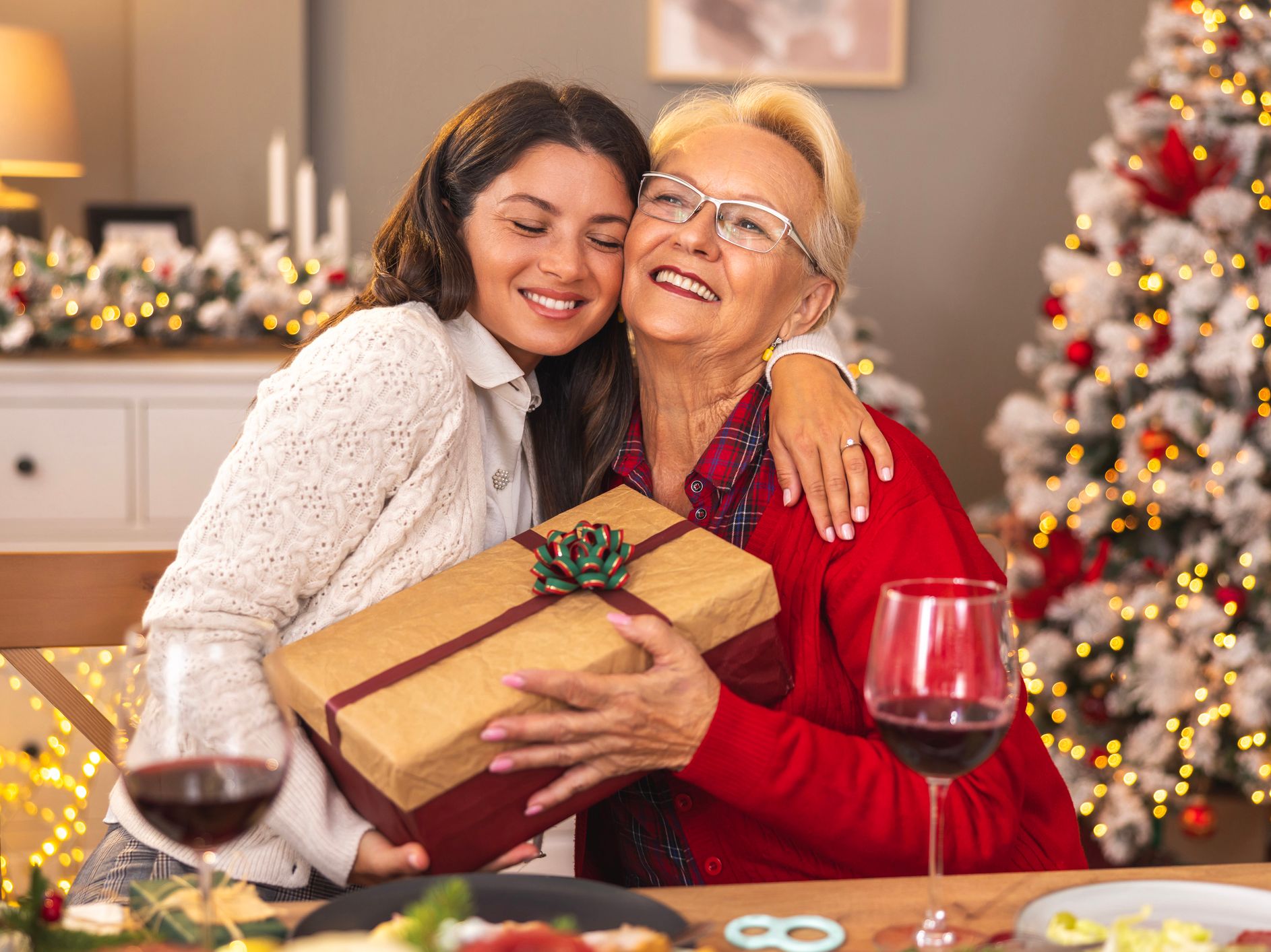 A young woman hugs her smiling grandmother while presenting a beautifully wrapped gift, with a glowing Christmas tree in the background, showcasing heartfelt Christmas gift ideas.