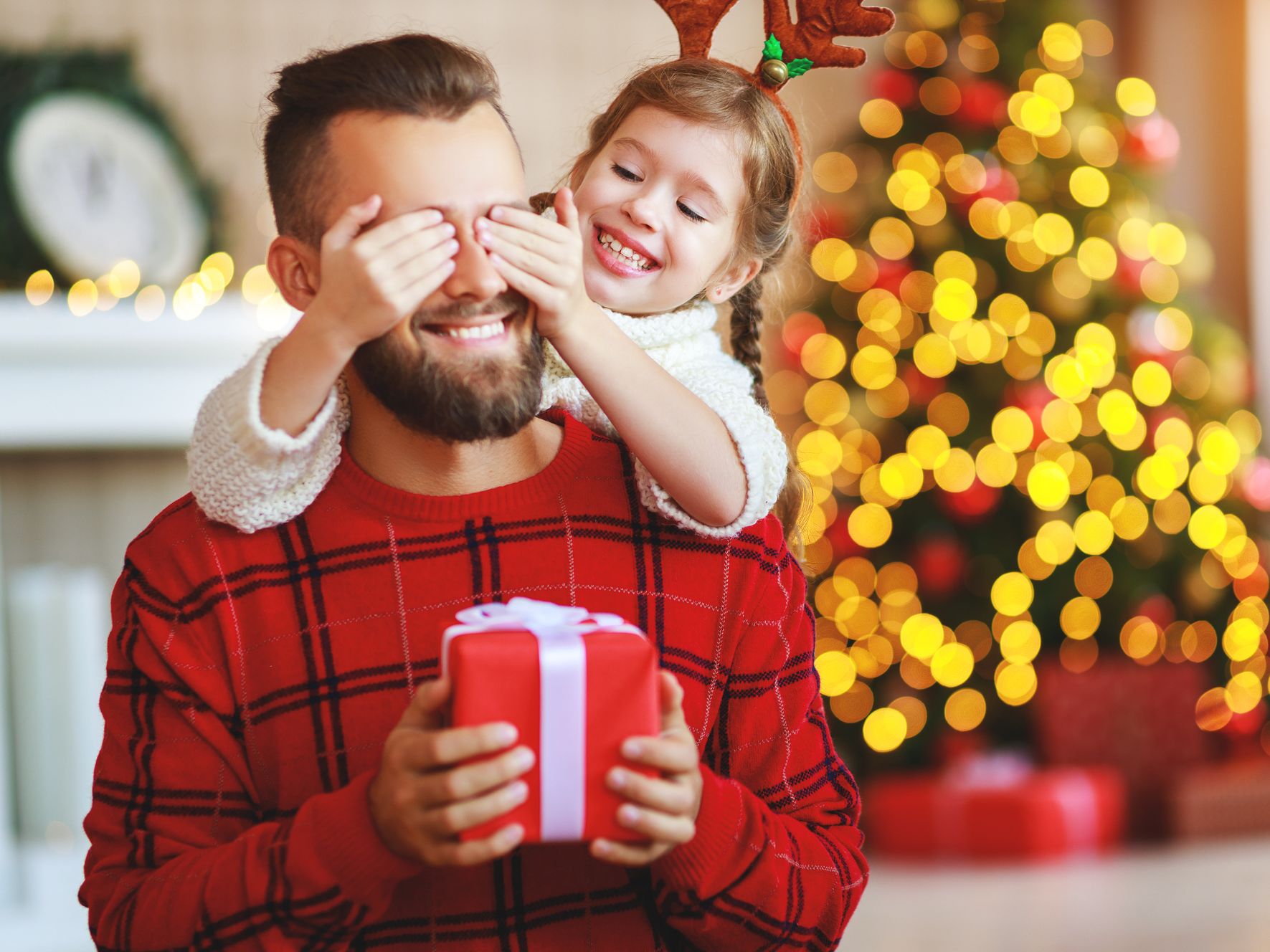 A little girl playfully covers her father's eyes as he holds a wrapped present, with a glowing Christmas tree in the background, showcasing joyful Christmas gift ideas.