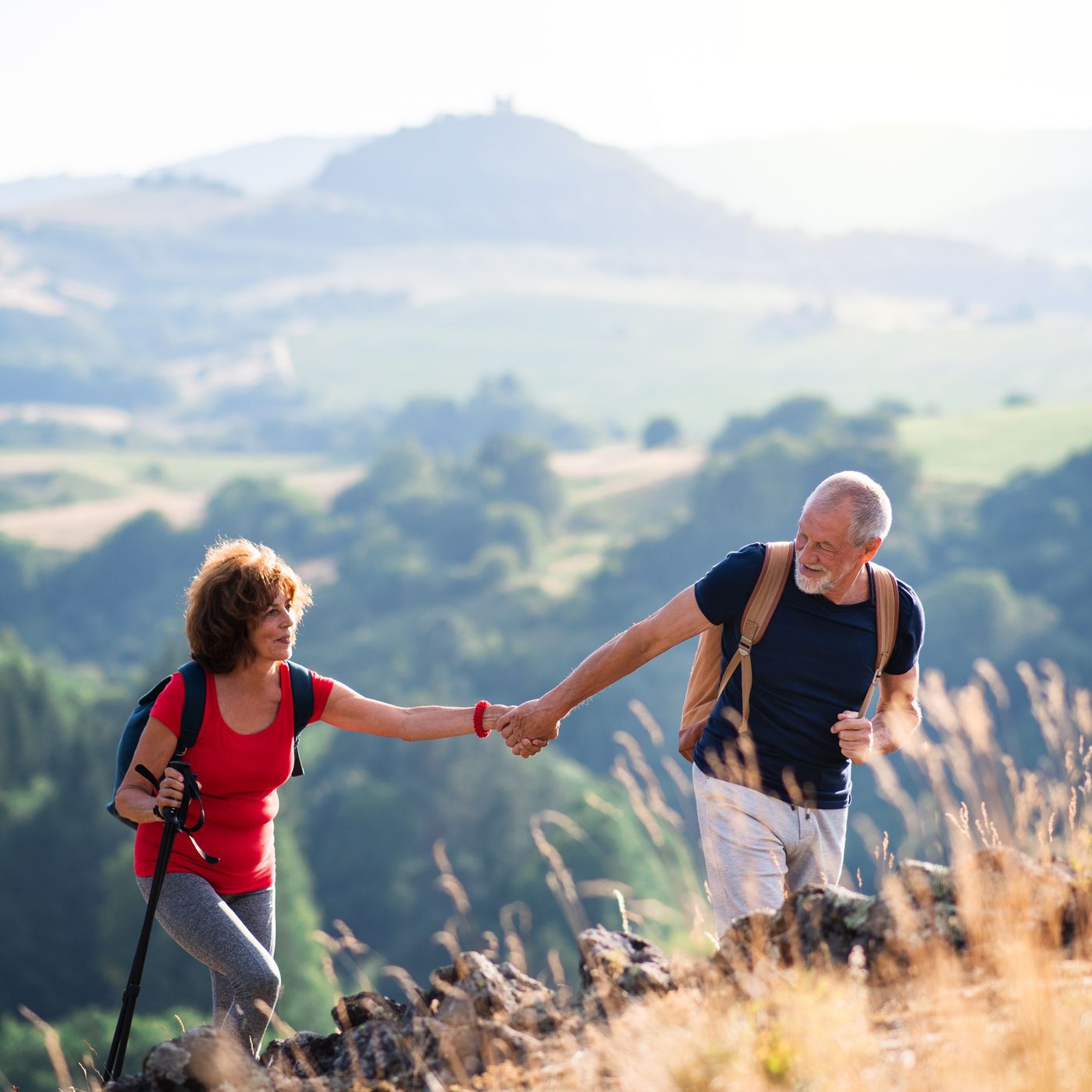 An active senior couple hiking in a scenic countryside, with the man helping the woman up a rocky hill. Rolling green hills and a soft, sunny backdrop enhance the view.