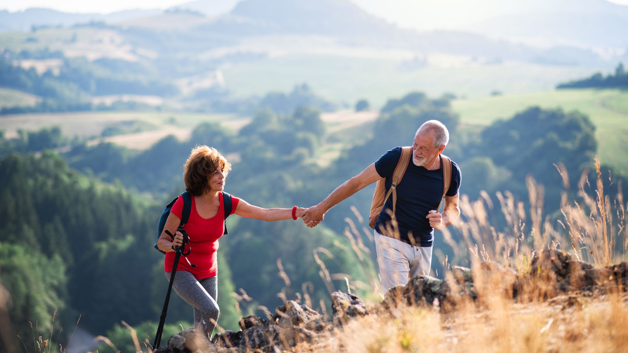 An active senior couple hiking in a scenic countryside, with the man helping the woman up a rocky hill. Rolling green hills and a soft, sunny backdrop enhance the view.