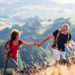 An active senior couple hiking in a scenic countryside, with the man helping the woman up a rocky hill. Rolling green hills and a soft, sunny backdrop enhance the view.