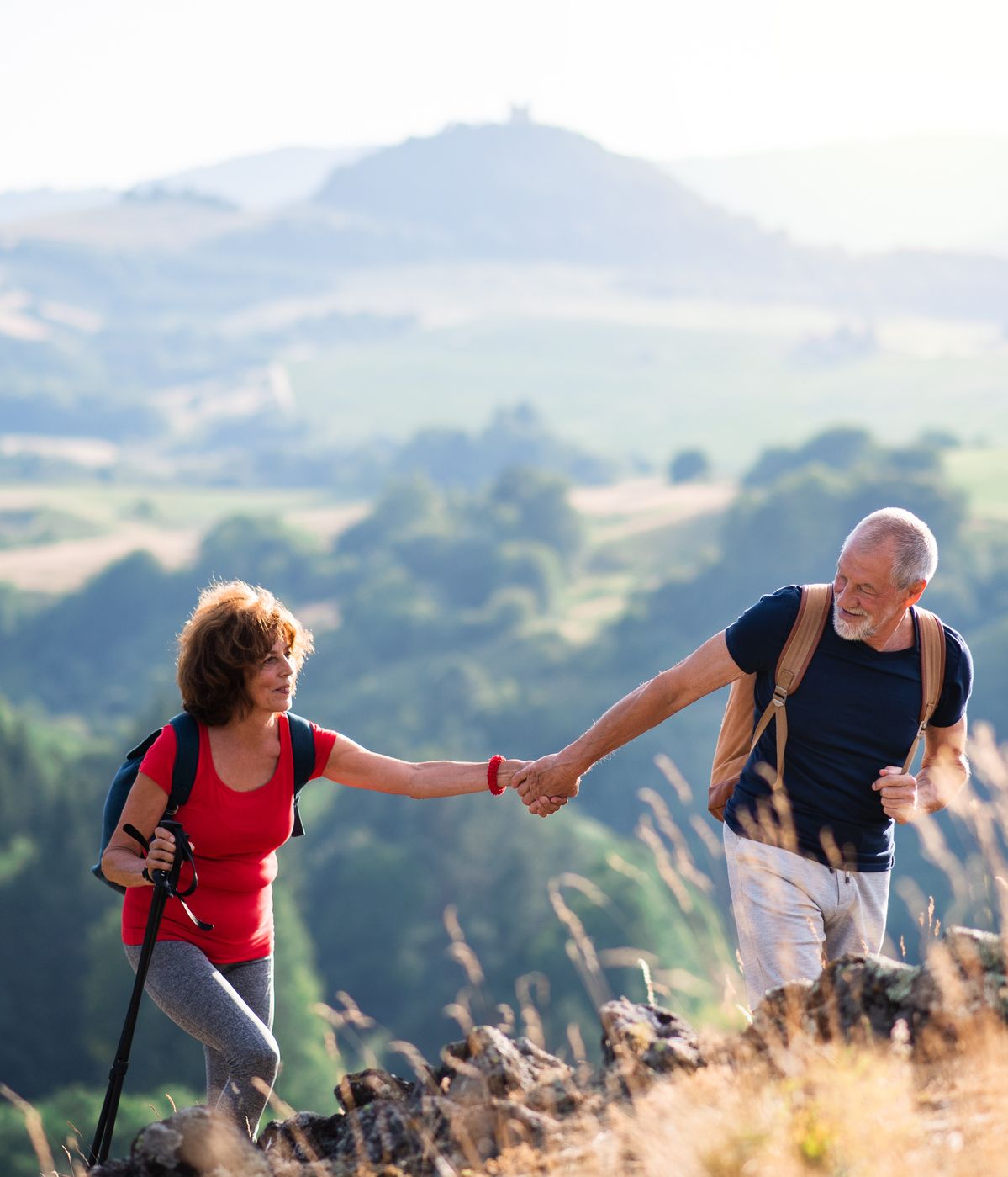 An active senior couple hiking in a scenic countryside, with the man helping the woman up a rocky hill. Rolling green hills and a soft, sunny backdrop enhance the view.