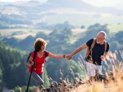 An active senior couple hiking in a scenic countryside, with the man helping the woman up a rocky hill. Rolling green hills and a soft, sunny backdrop enhance the view.