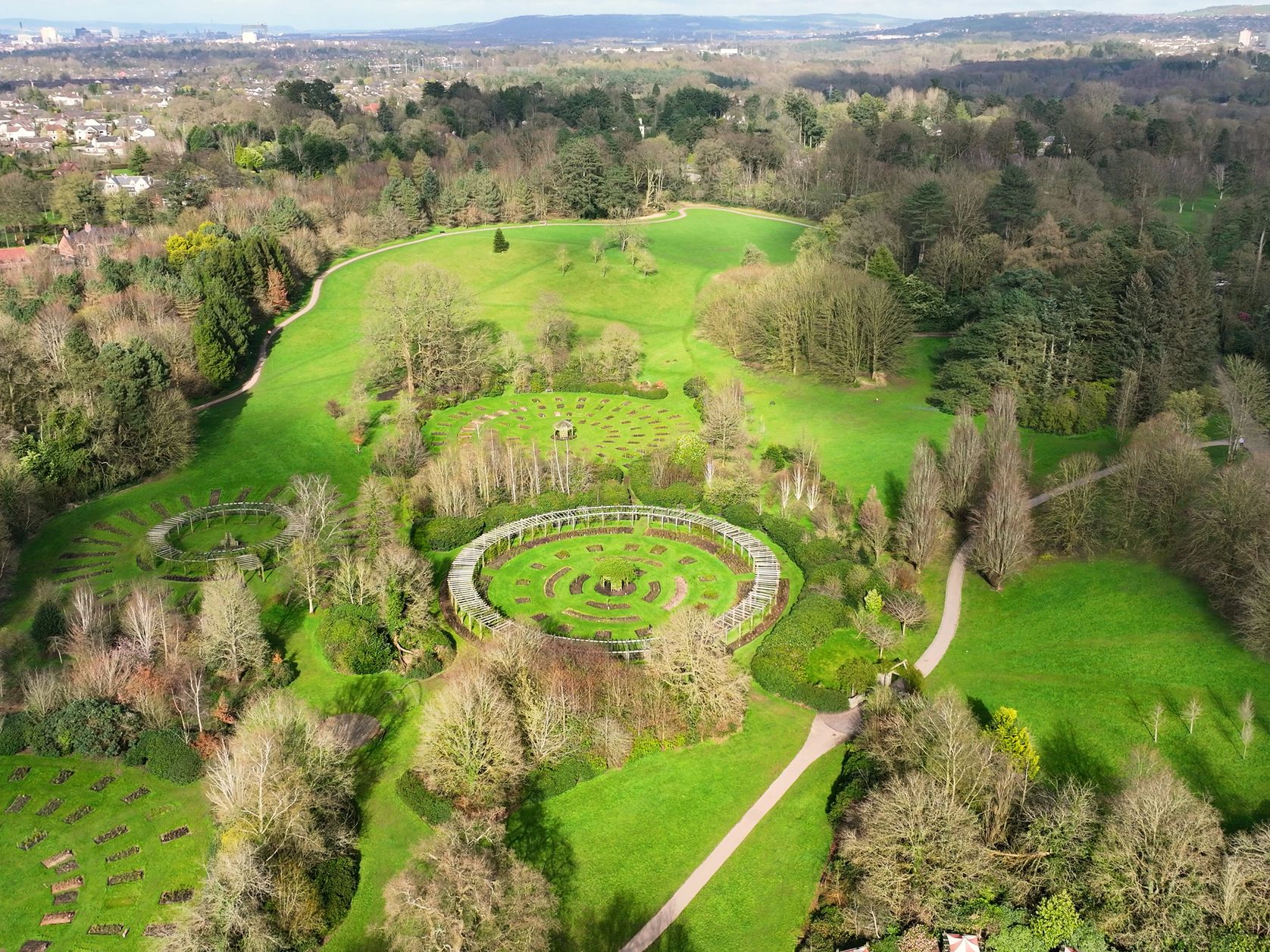 An aerial view of a large park with lush green lawns, circular garden arrangements, walking paths, and surrounding trees, with a distant town visible on the horizon.