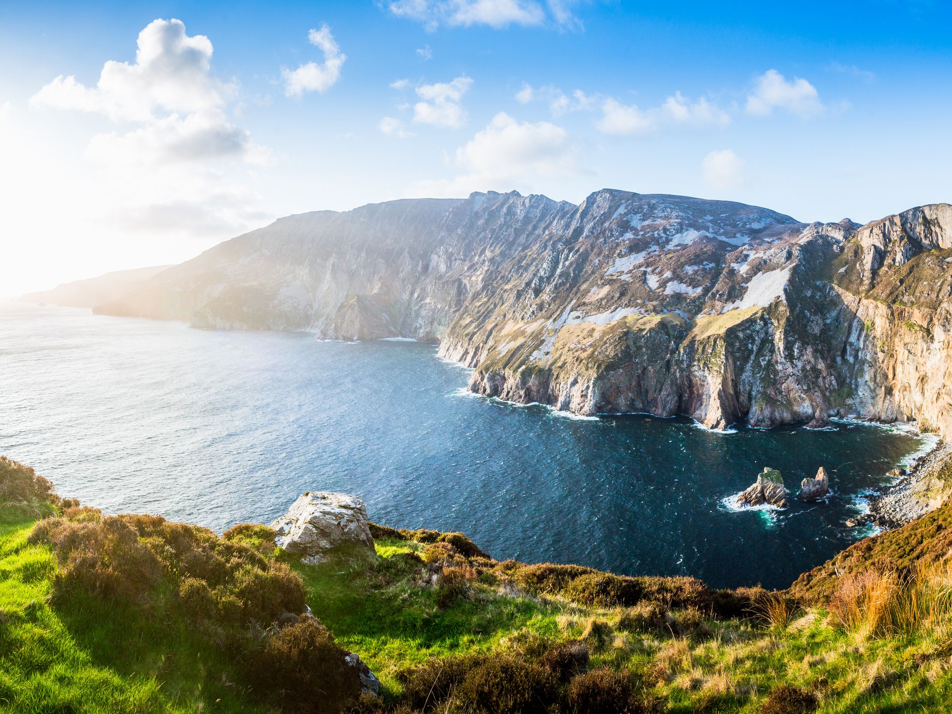 A stunning coastal view of towering cliffs meeting the deep blue ocean, with grassy hills in the foreground and sunlight illuminating the scene under a bright, partly cloudy sky.
