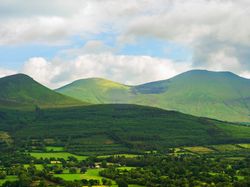 A lush, green landscape featuring rolling fields and dense forests, with three towering, grass-covered hills in the background under a partly cloudy blue sky.