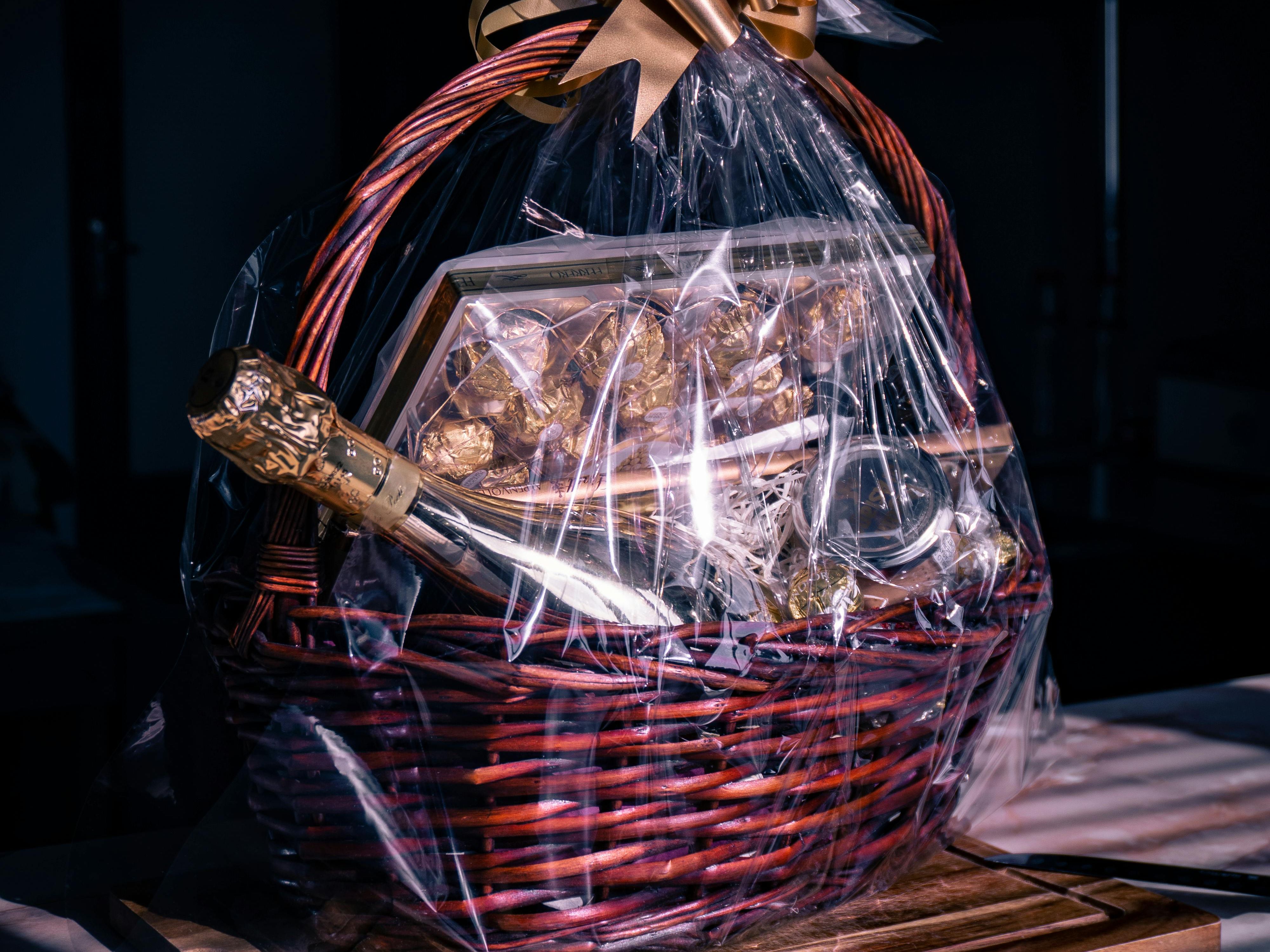 A luxurious gift basket wrapped in clear cellophane with a gold ribbon, containing a bottle of champagne, gourmet chocolates, and elegant treats, placed on a wooden board.