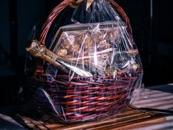A luxurious gift basket wrapped in clear cellophane with a gold ribbon, containing a bottle of champagne, gourmet chocolates, and elegant treats, placed on a wooden board.