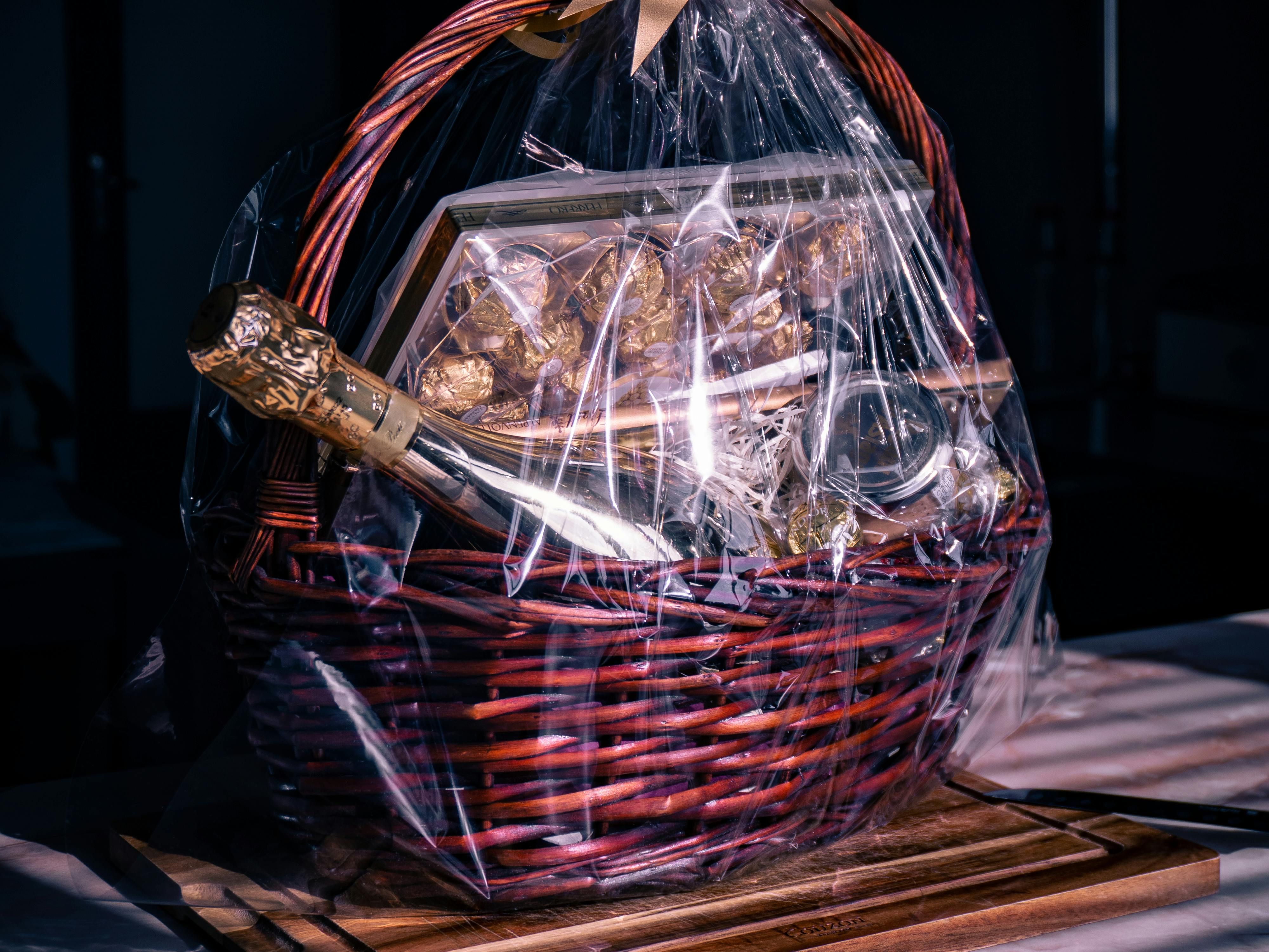 A luxurious gift basket wrapped in clear cellophane with a gold ribbon, containing a bottle of champagne, gourmet chocolates, and elegant treats, placed on a wooden board.