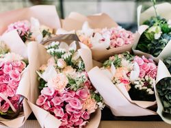 A collection of beautifully arranged flower bouquets wrapped in soft pastel paper, featuring pink roses, hydrangeas, carnations, and white lilies with lush green foliage.
