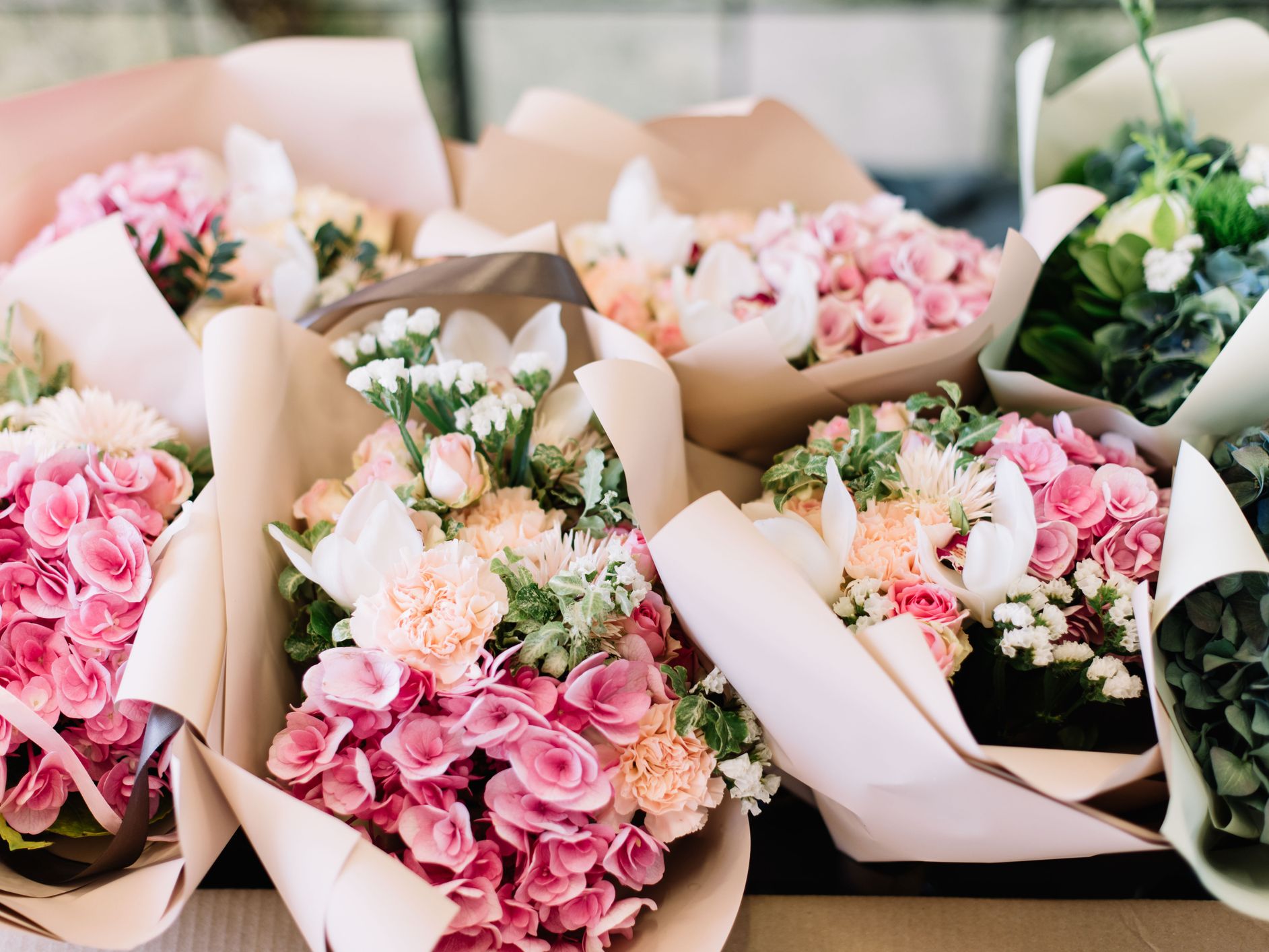 A collection of beautifully arranged flower bouquets wrapped in soft pastel paper, featuring pink roses, hydrangeas, carnations, and white lilies with lush green foliage.