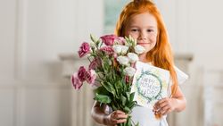 A young girl with long red hair smiles while holding a bouquet of pink and white flowers and a handmade 