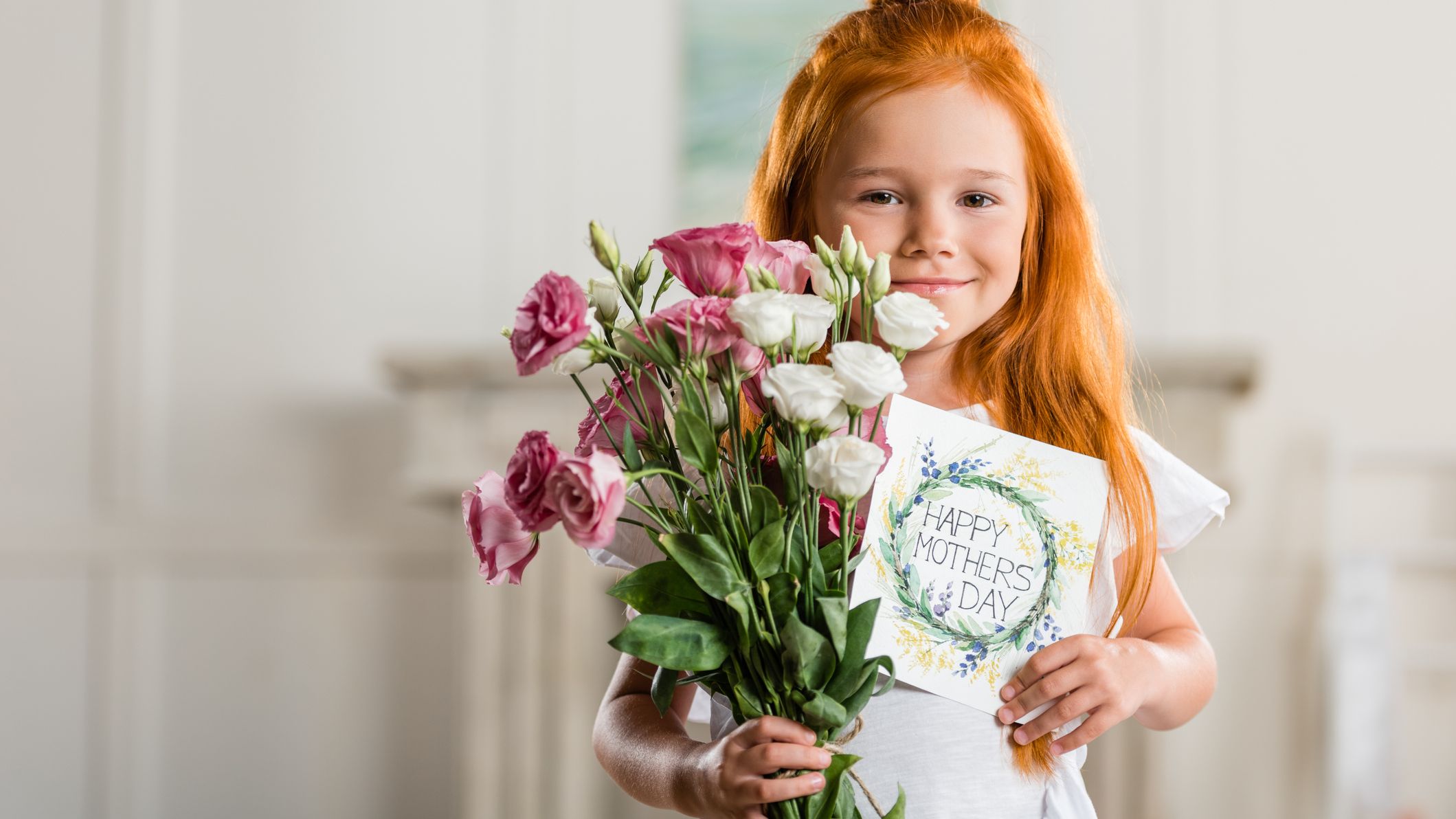 A young girl with long red hair smiles while holding a bouquet of pink and white flowers and a handmade 