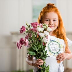 A young girl with long red hair smiles while holding a bouquet of pink and white flowers and a handmade 