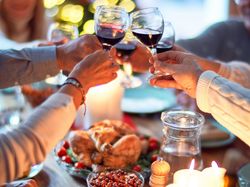 Group of people clinking wine glasses around a festive dinner table with candles and food, in front of a lit Christmas tree.