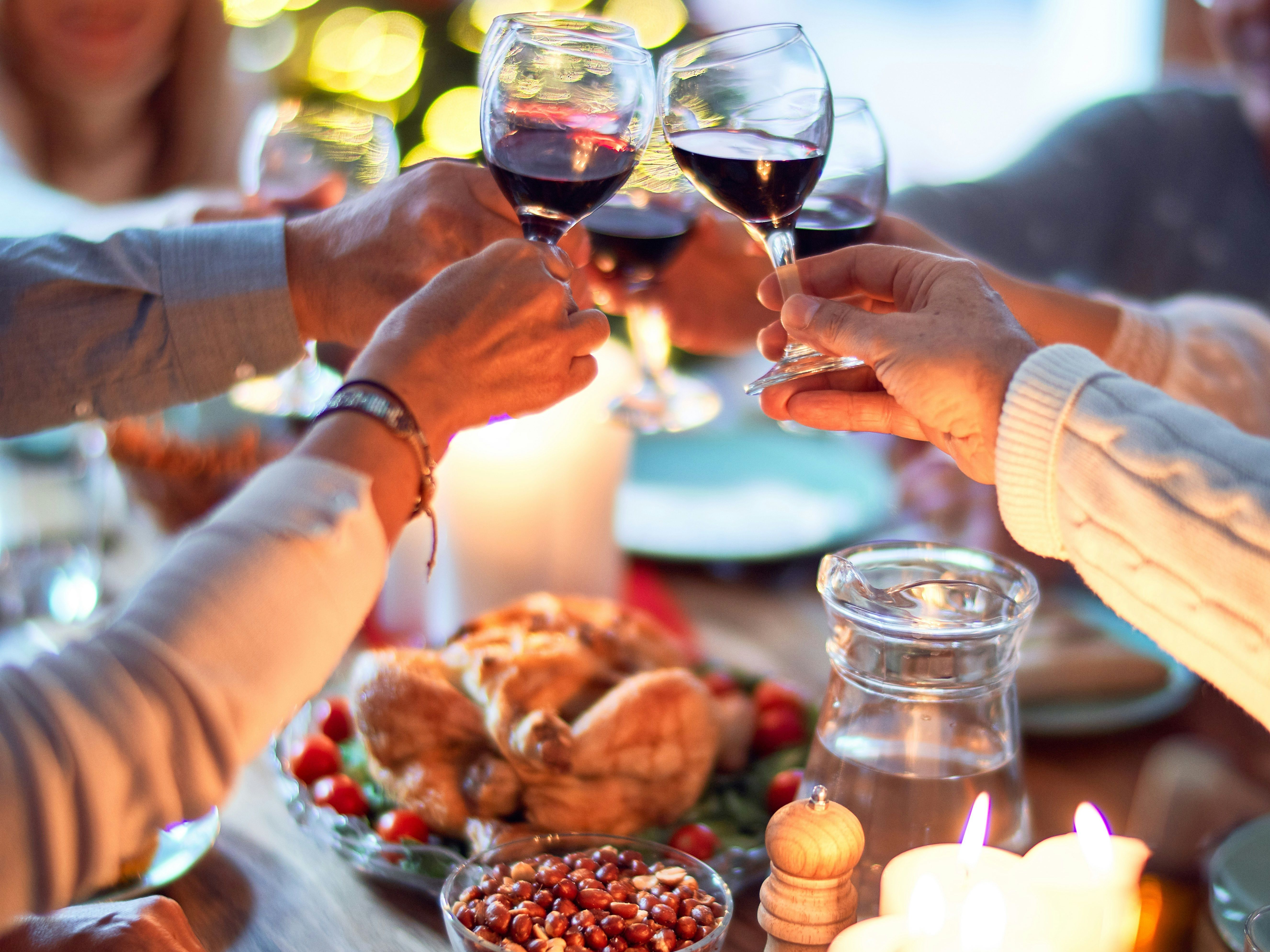 Group of people clinking wine glasses around a festive dinner table with candles and food, in front of a lit Christmas tree.