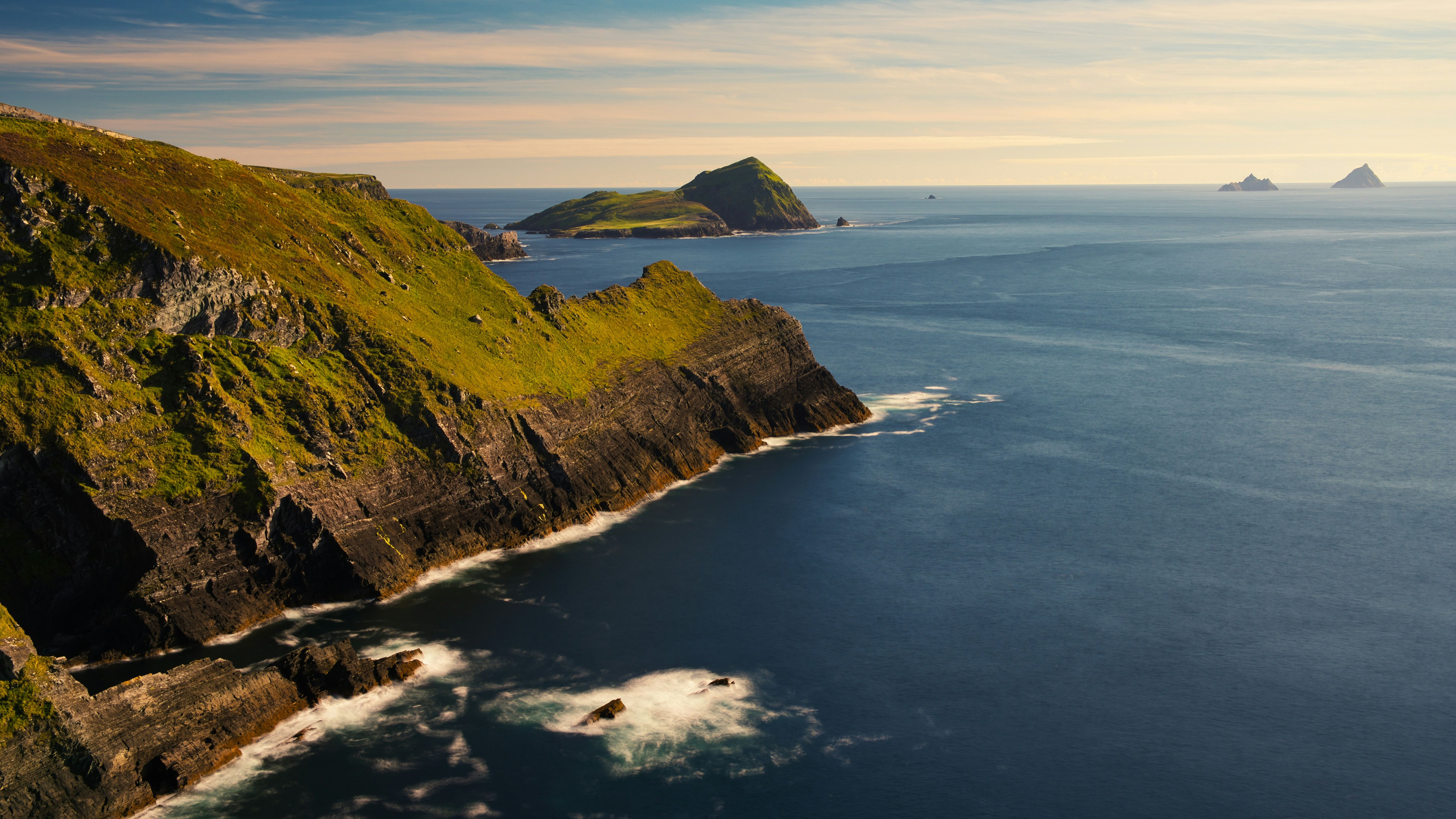Dramatic coastal cliffs with green hills overlooking the ocean, with islands visible in the distance under a partly cloudy sky.