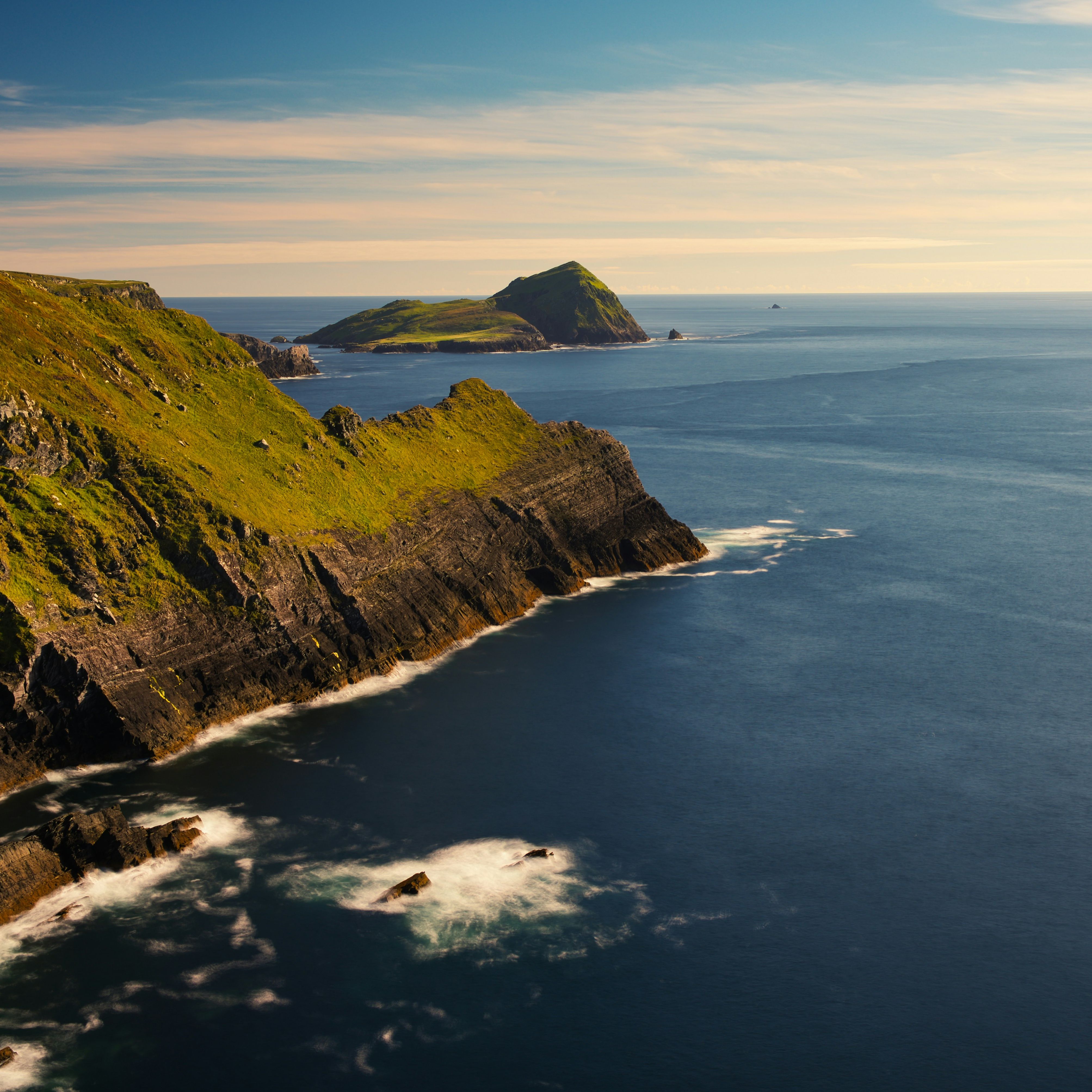 Dramatic coastal cliffs with green hills overlooking the ocean, with islands visible in the distance under a partly cloudy sky.