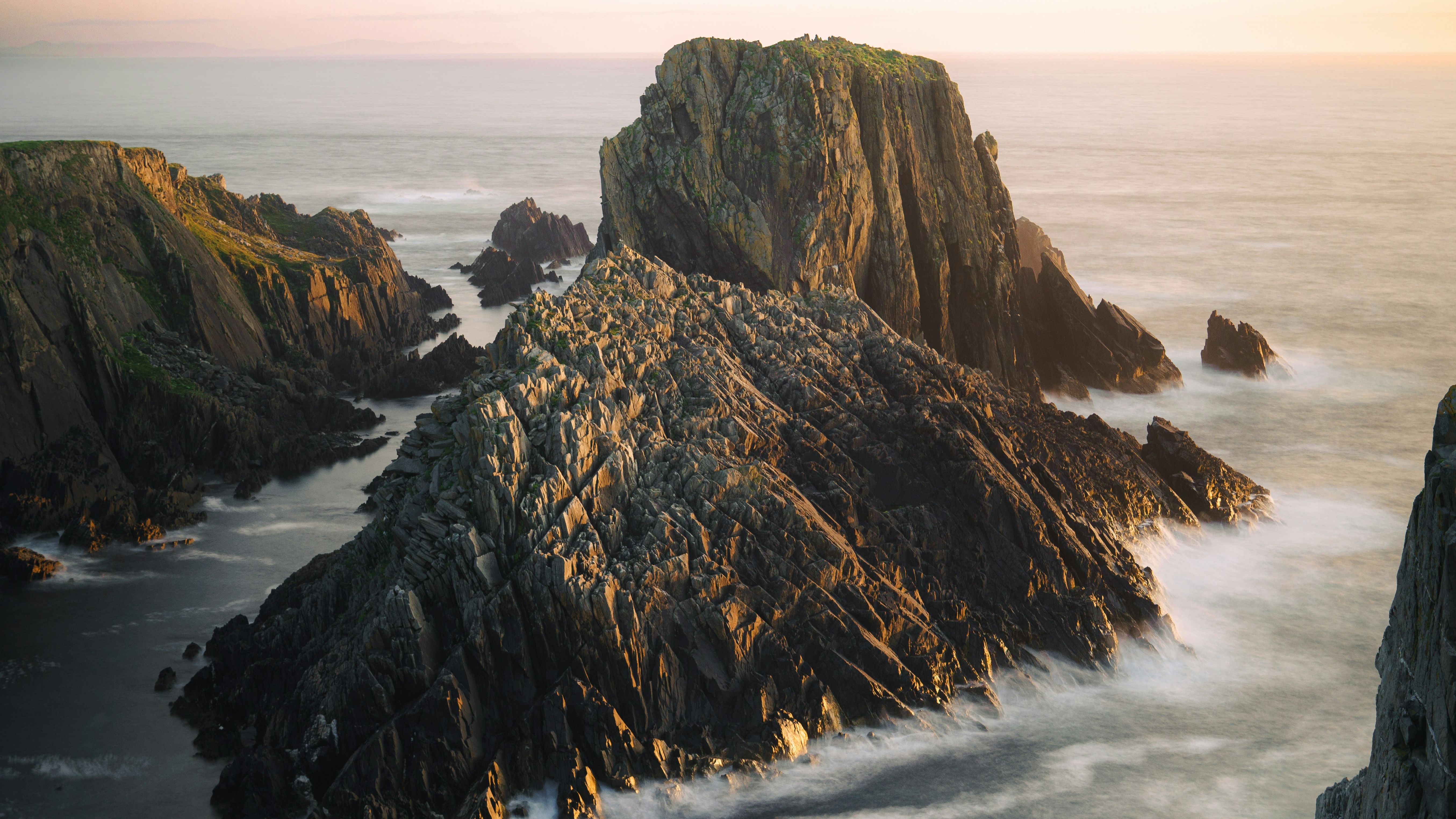 Jagged rocky cliffs rising from the sea at sunset with waves crashing against the rocks.