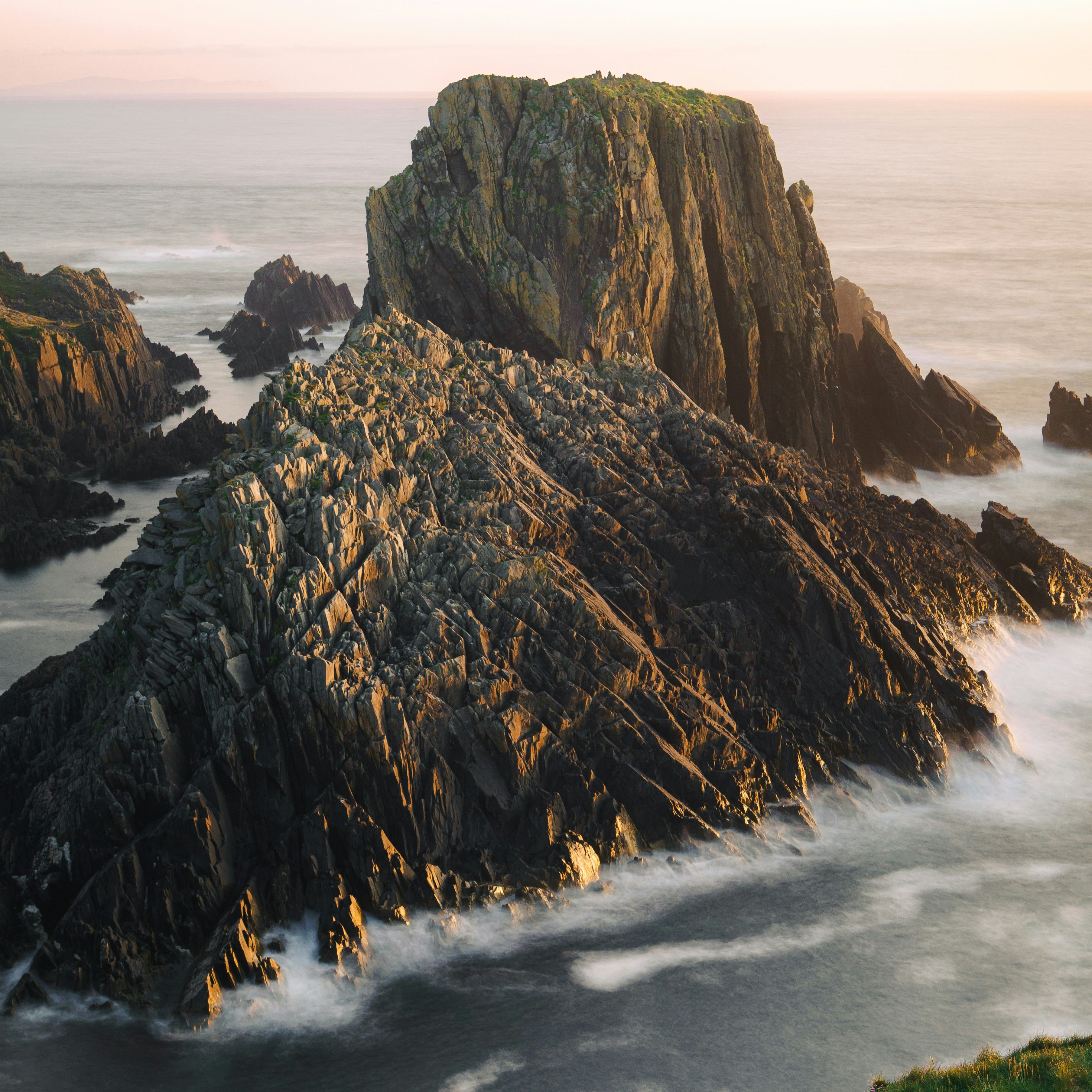 Jagged rocky cliffs rising from the sea at sunset with waves crashing against the rocks.