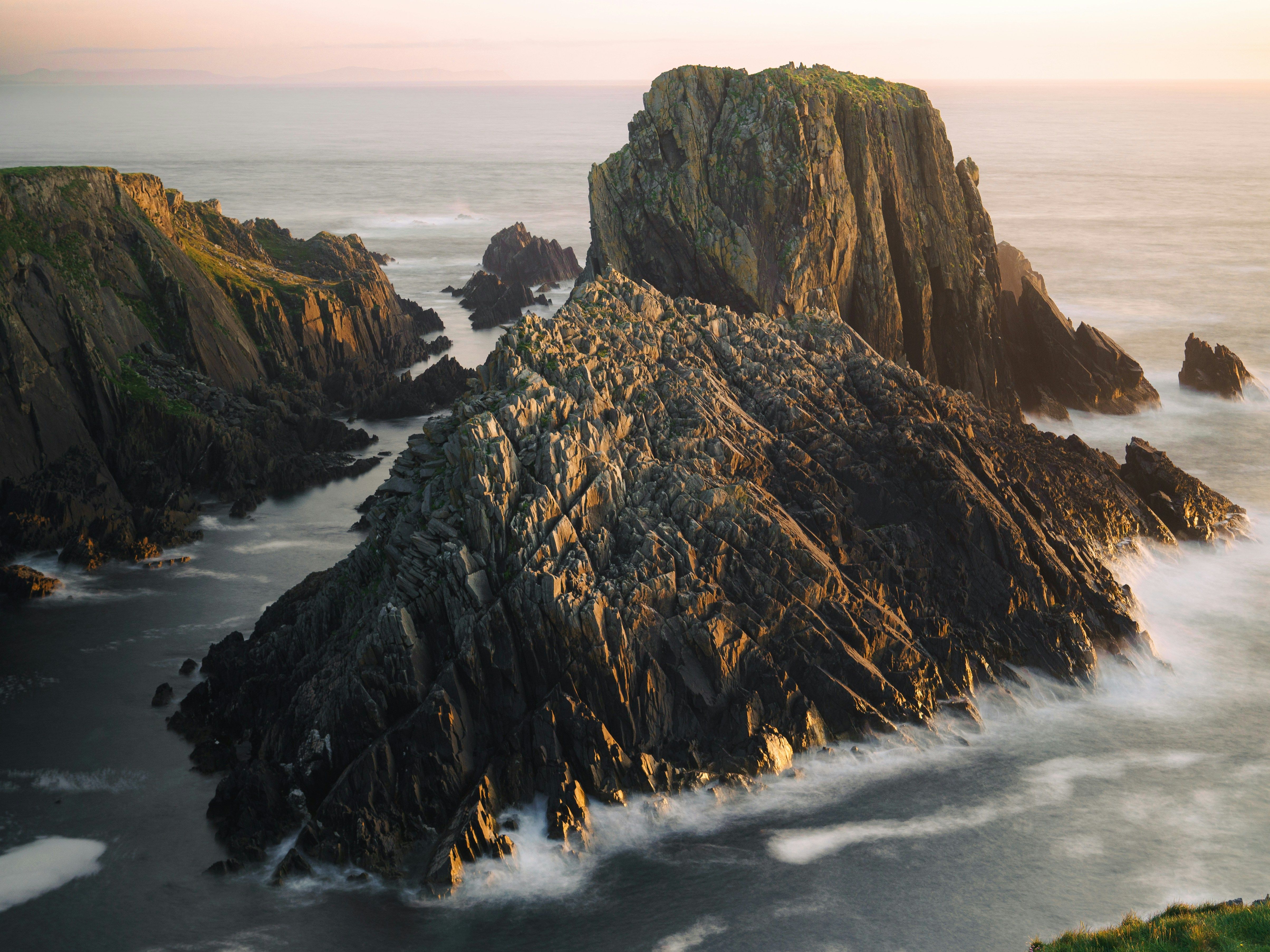 Jagged rocky cliffs rising from the sea at sunset with waves crashing against the rocks.