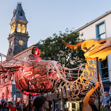 Illuminated bird skeleton street puppet carried in a parade in front of a clock tower at dusk