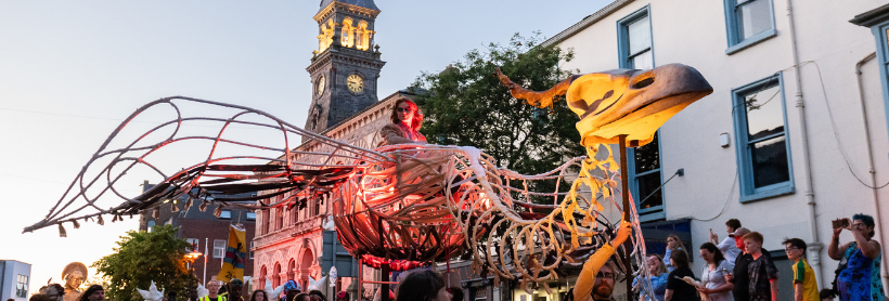 Illuminated bird skeleton street puppet carried in a parade in front of a clock tower at dusk