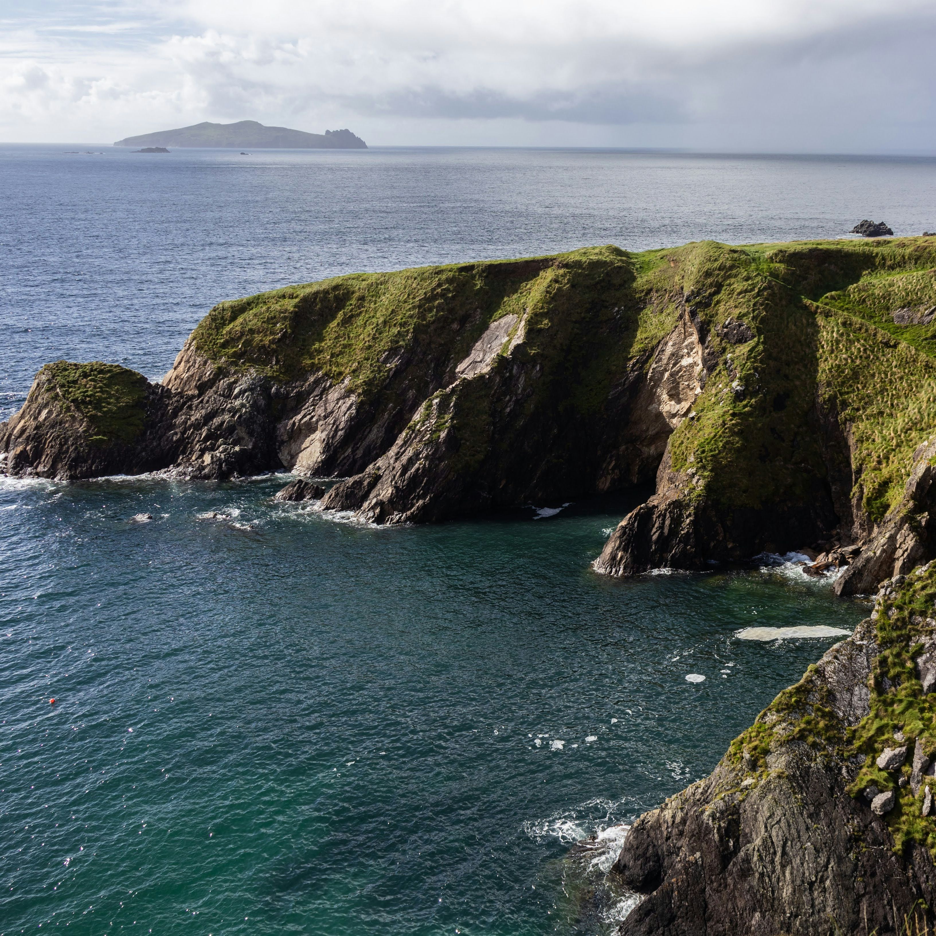 Rugged grassy sea cliffs above a calm bay with an island on the horizon under cloudy skies