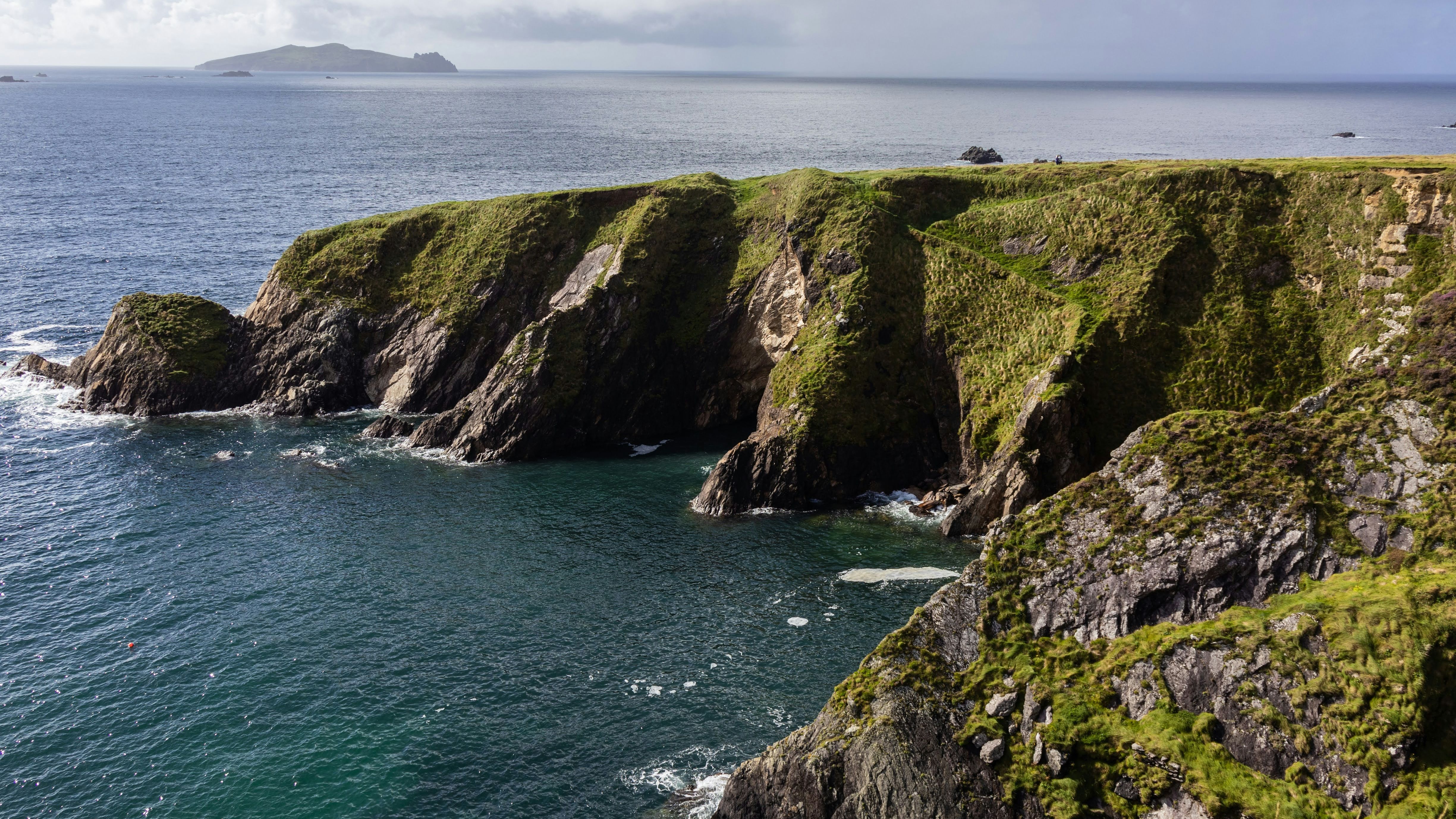 Rugged grassy sea cliffs above a calm bay with an island on the horizon under cloudy skies
