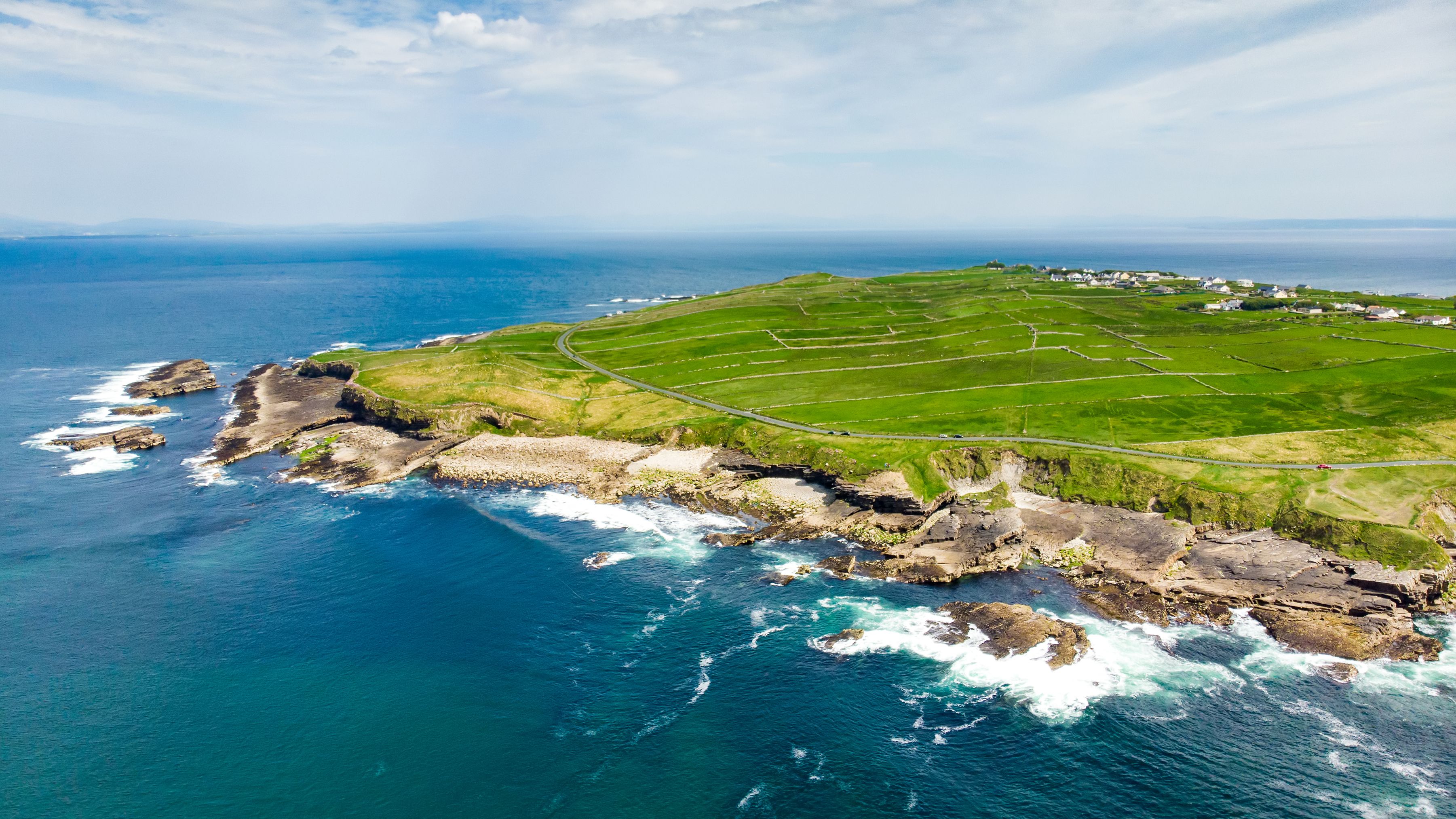 Aerial view of a rugged coastline with green fields, a small village and waves breaking on rocky cliffs under a bright, cloudy sky