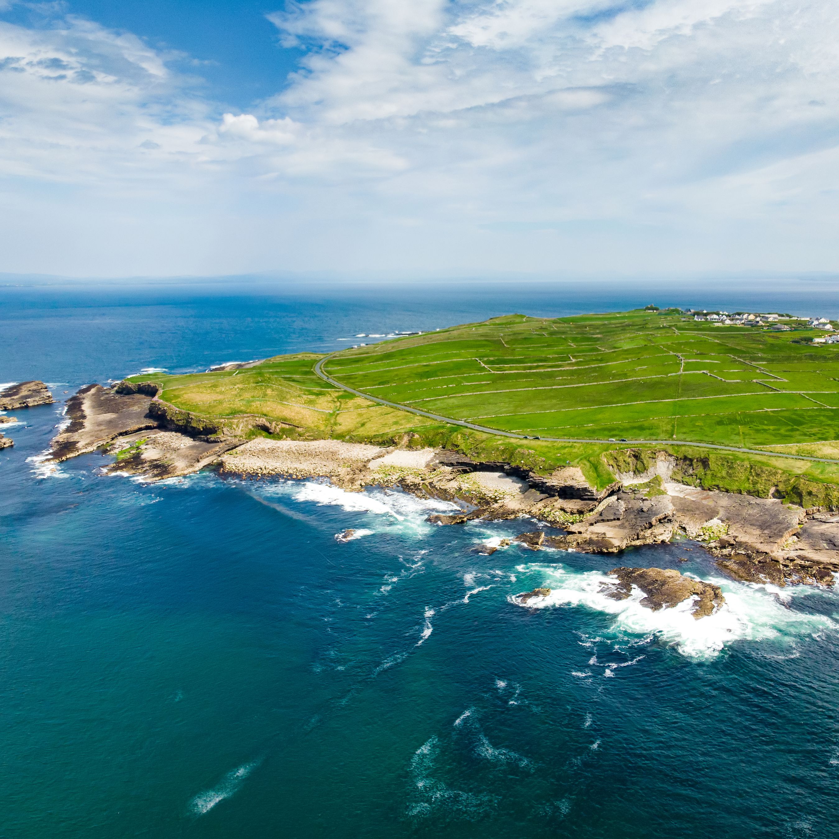 Aerial view of a rugged coastline with green fields, a small village and waves breaking on rocky cliffs under a bright, cloudy sky