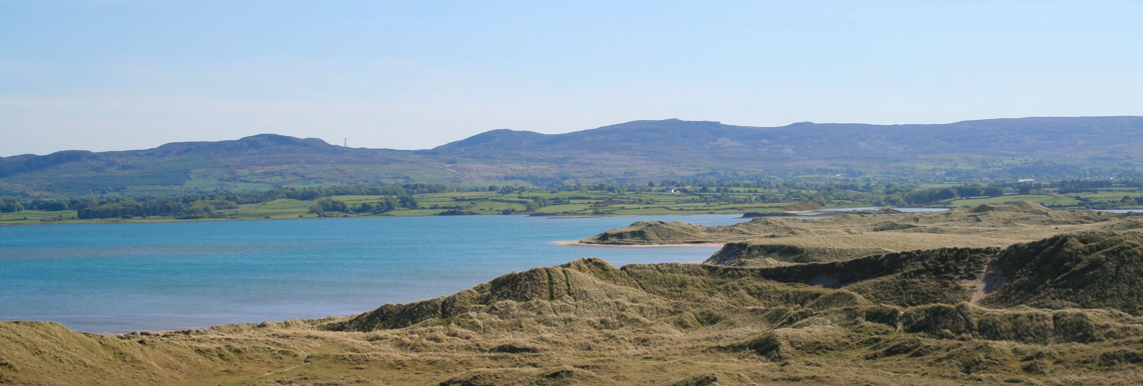 Coastal sand dunes overlooking a calm blue bay with green hills in the distance