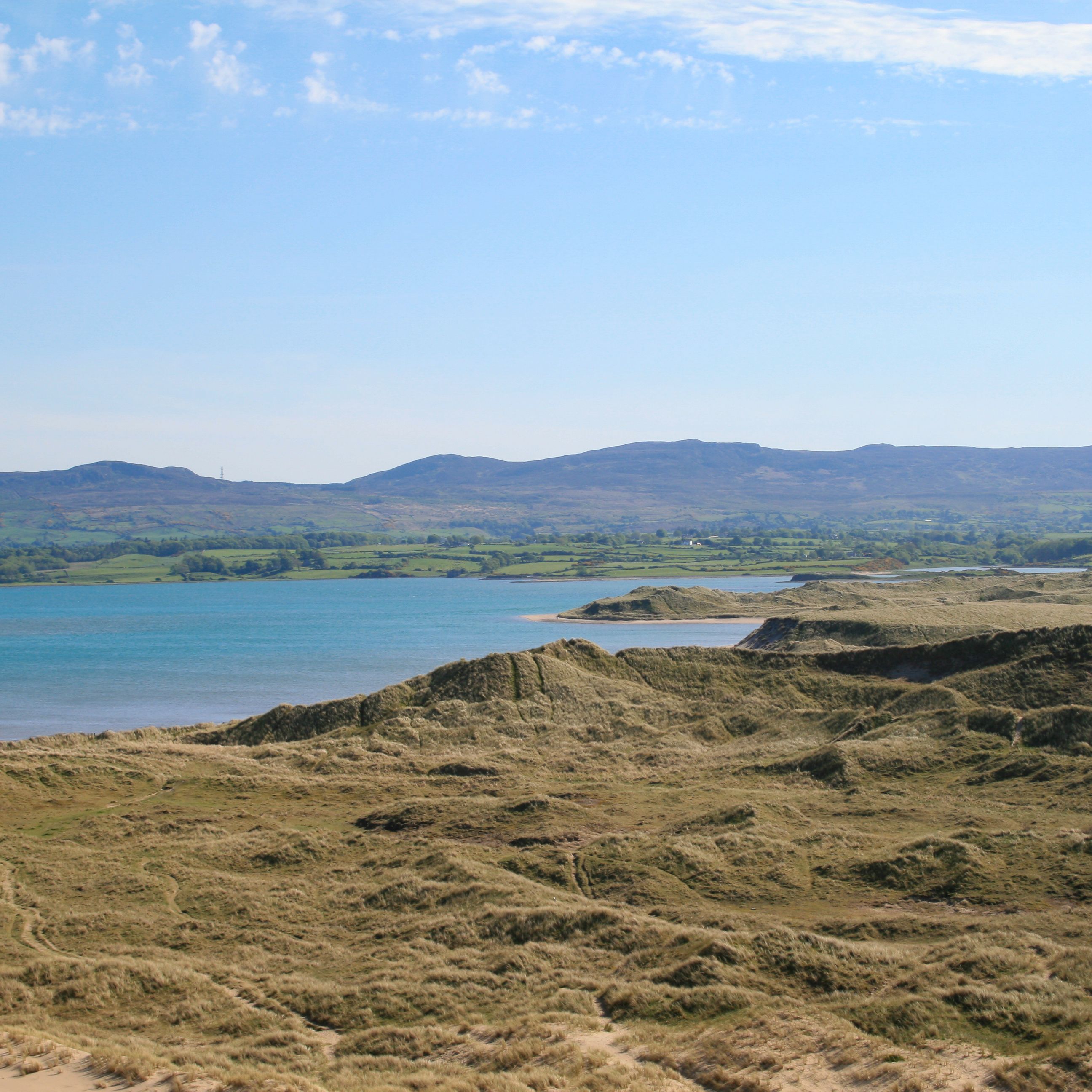 Coastal sand dunes overlooking a calm blue bay with green hills in the distance