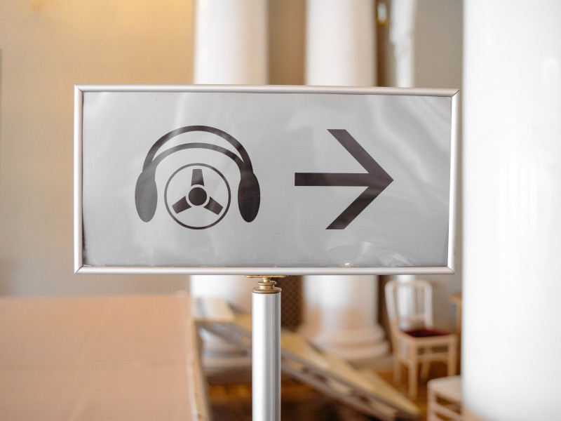 A sign with an icon of headphones and an audio guide symbol, followed by a right-pointing arrow, indicating the direction to an audio tour or guide service.