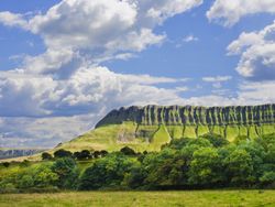 The beautiful Benbulben landscape with a unique, flat top and green, terraced slopes. Below, a mix of forest and fields provides a serene foreground under a partly cloudy sky.