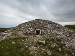A large, weathered stone cairn with an entrance opening at the base, set in a grassy, remote landscape. The overcast sky adds a somber tone to this ancient monument.