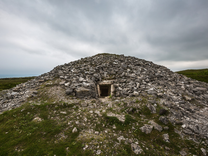 A large, weathered stone cairn with an entrance opening at the base, set in a grassy, remote landscape. The overcast sky adds a somber tone to this ancient monument.