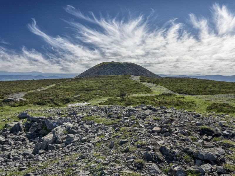 A stone-covered cairn atop a hill, with a winding path leading to it. The sky is filled with streaking clouds, creating a dynamic backdrop against the vast open landscape.